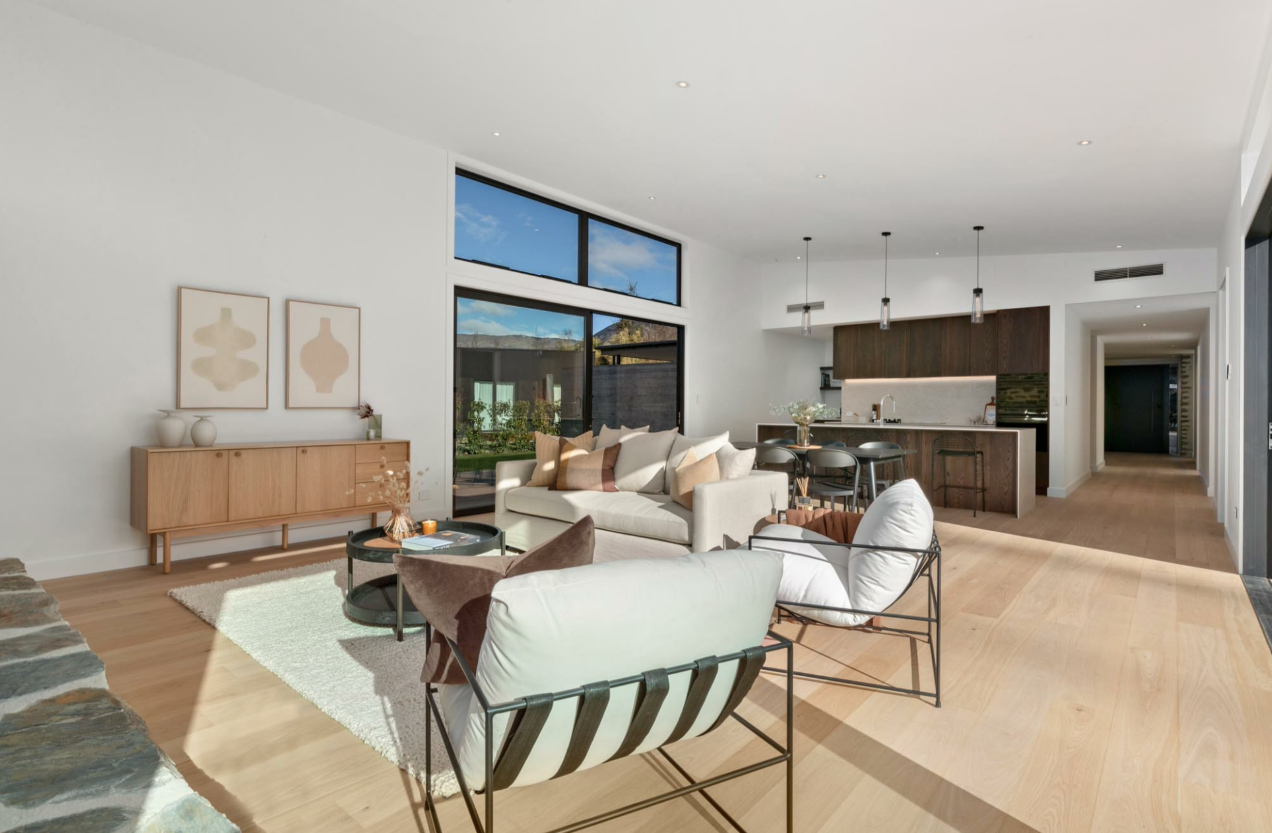 Interior view of a modern, open-concept living room with high ceilings, large windows, and minimalist decor. Features include a white sofa, armchairs, a black coffee table, a light wood sideboard with decorative vases, and abstract wall art. The space opens to a kitchen with dark wood cabinets, pendant lighting, and a dining area.