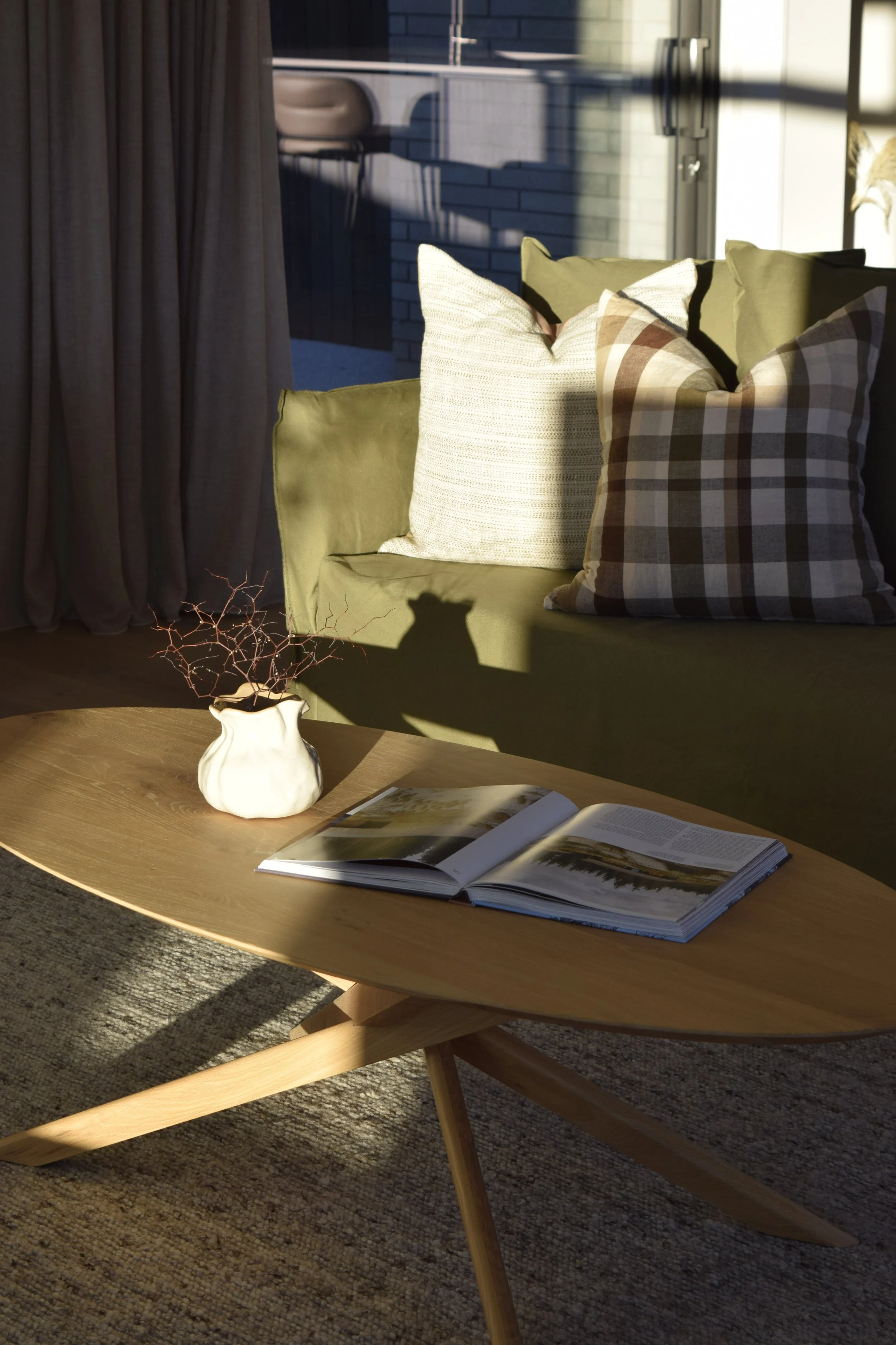 A coffee table with an open magazine and a small white vase with twigs inside. In the background, there is a green couch with patterned pillows, sunlight casting shadows, a dark curtain on the left, and a kitchen area with high chairs and a fridge.