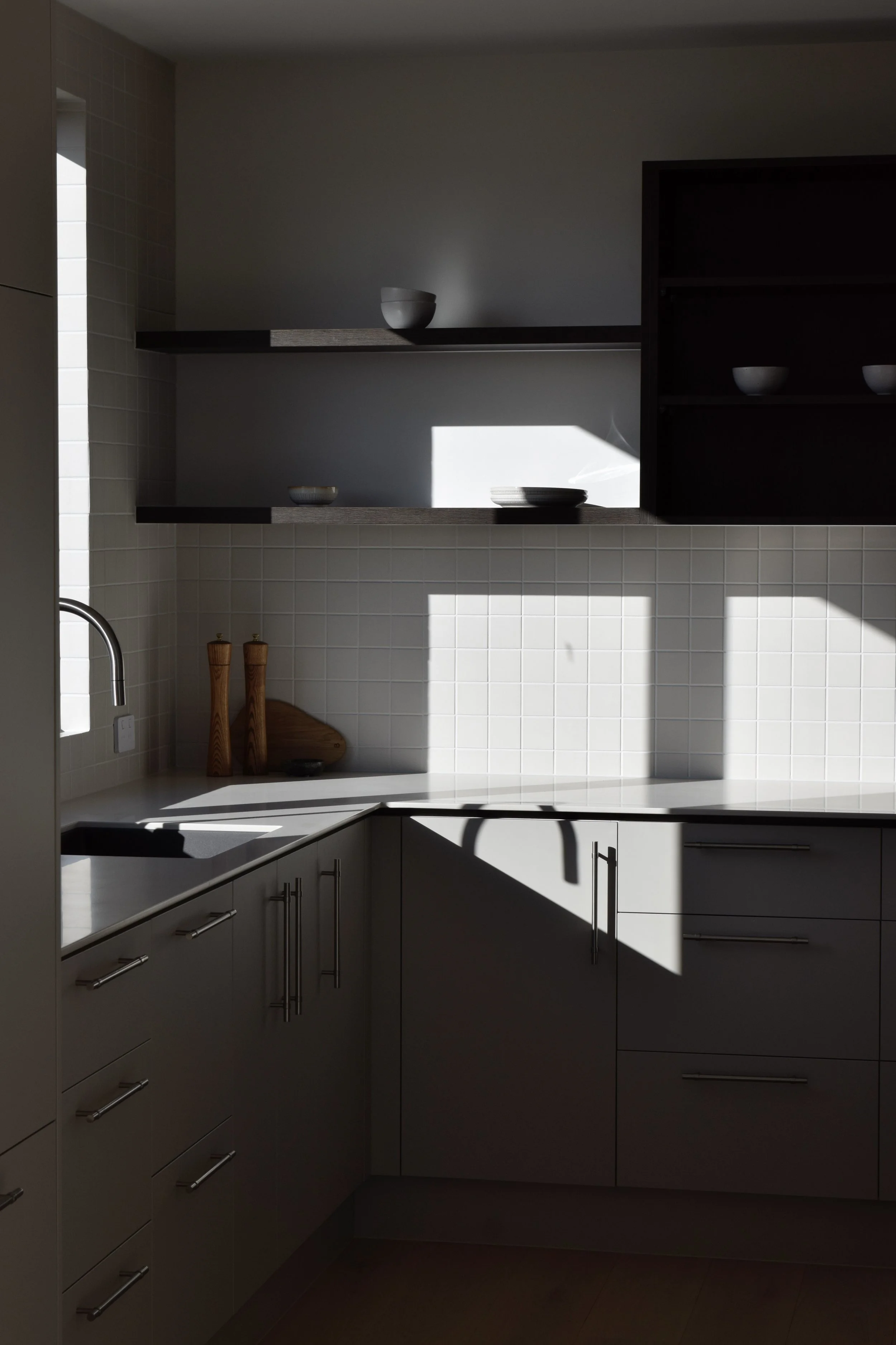 Minimalist kitchen with white cabinets, a white countertop, and white tiled walls. Open black shelves hold white bowls, with sunlight casting shadows on the wall.
