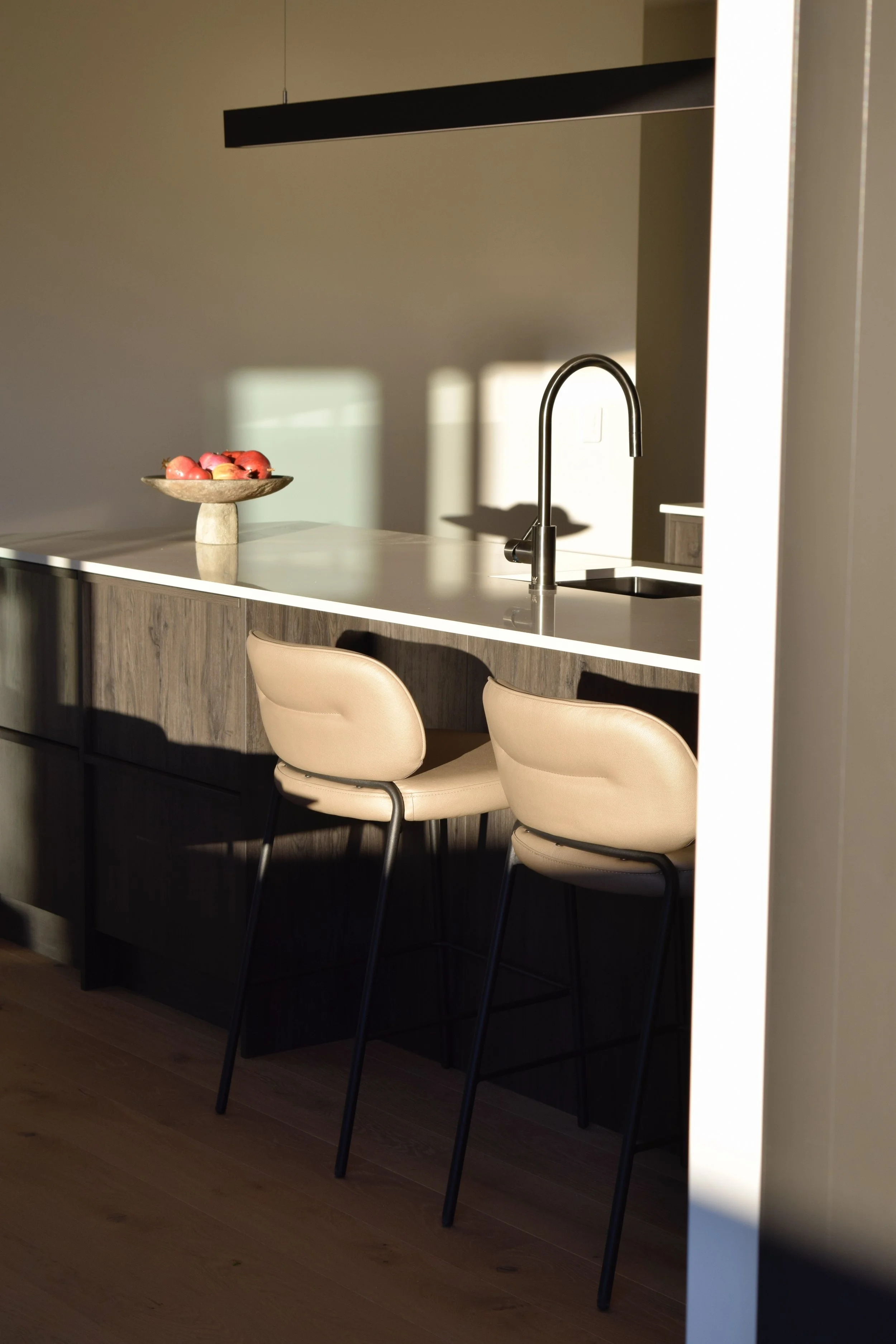 A modern kitchen with a white countertop, a black sink, and a curved faucet. There are two beige chairs with black legs in front of the counter. A decorative bowl of red and pink apples sits on the far left side of the counter. The kitchen has wood l