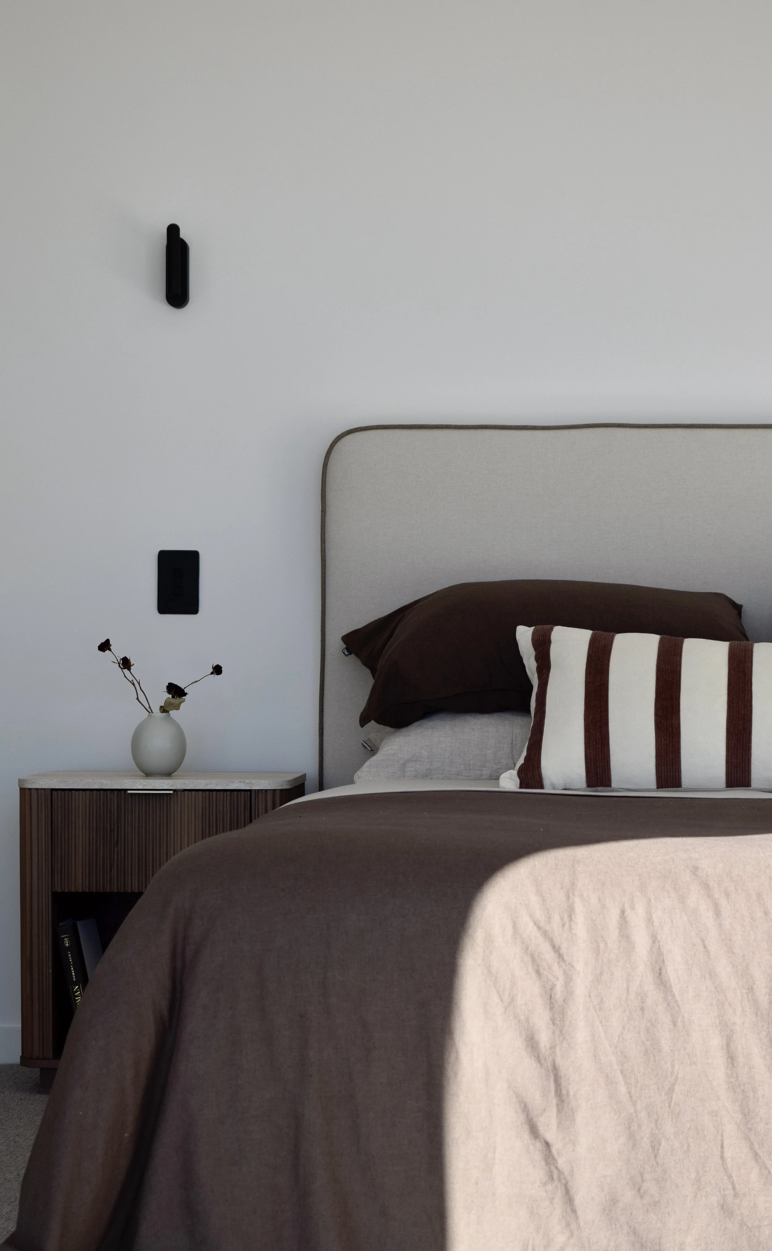 A bedroom with a bed, brown blanket, white pillow with brown stripes, beige headboard, and side table with a vase and dried flowers, in front of a white wall with black wall-mounted lamp and outlet.