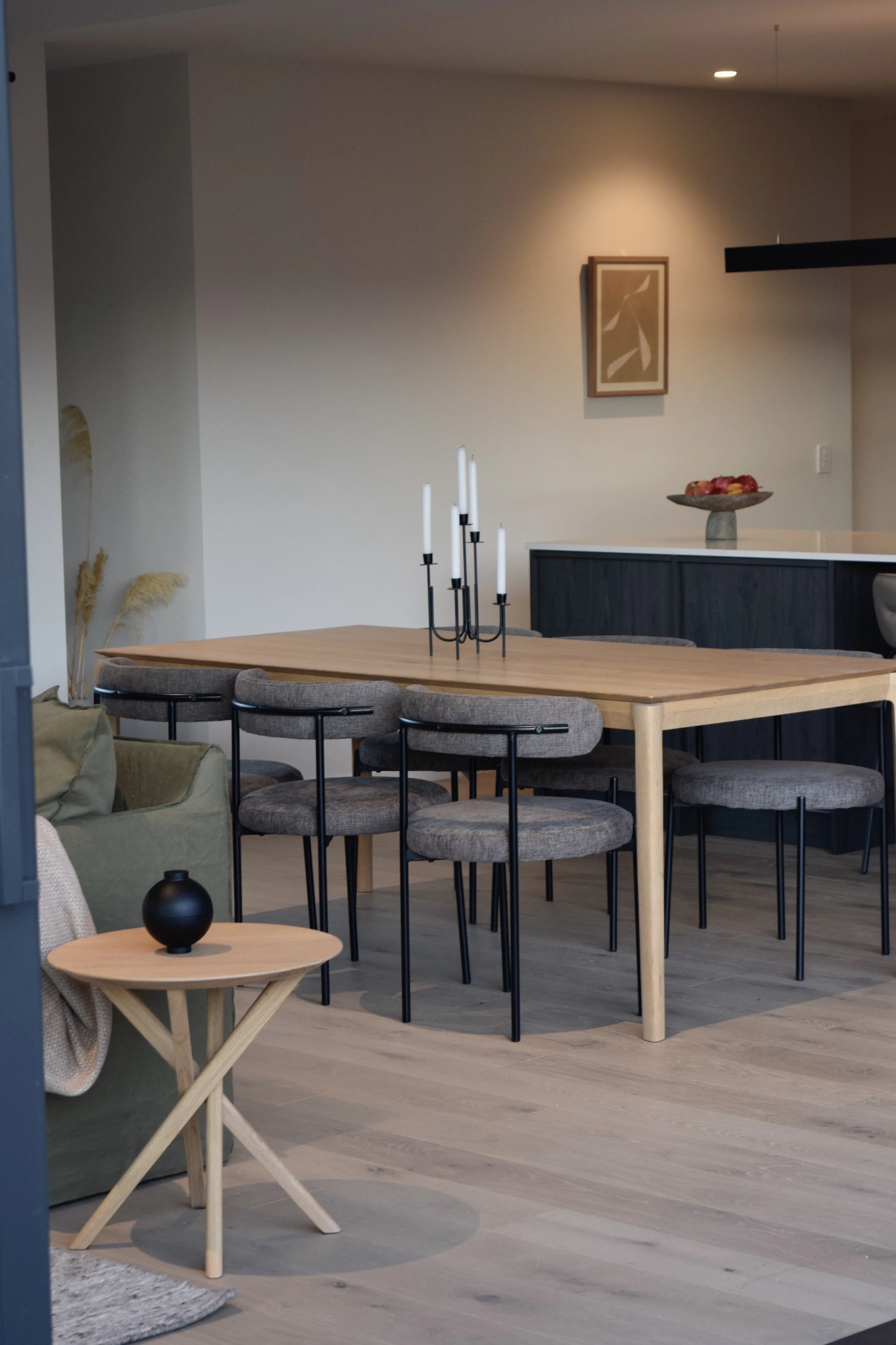 Modern dining area with a large wooden table, cushioned chairs, a candelabra, a small side table with a black vase, a decorative bowl of fruit on a black sideboard, and minimalist wall art illuminated by a wall-mounted light.