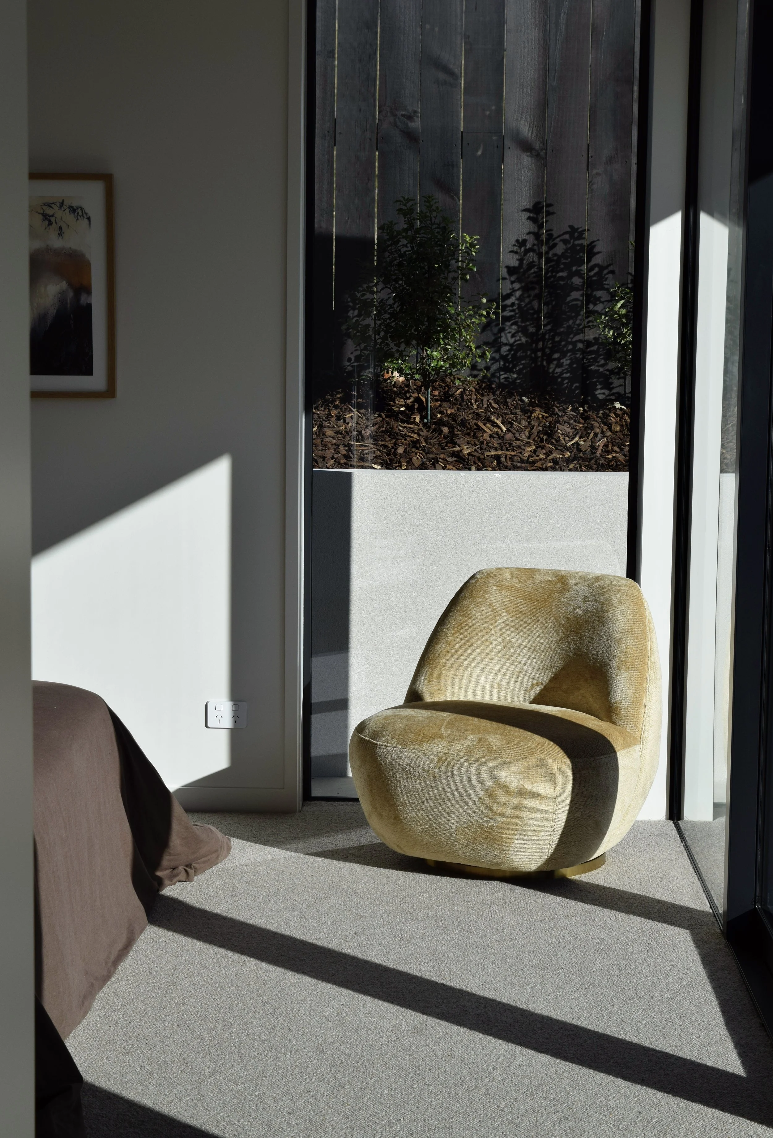 Interior of a room with cream carpet, a beige cozy chair, a partially visible brown sofa with a brown pillow, white walls, a framed landscape painting, and a sliding glass door leading outside to a small garden with mulch and a wooden privacy fence, 