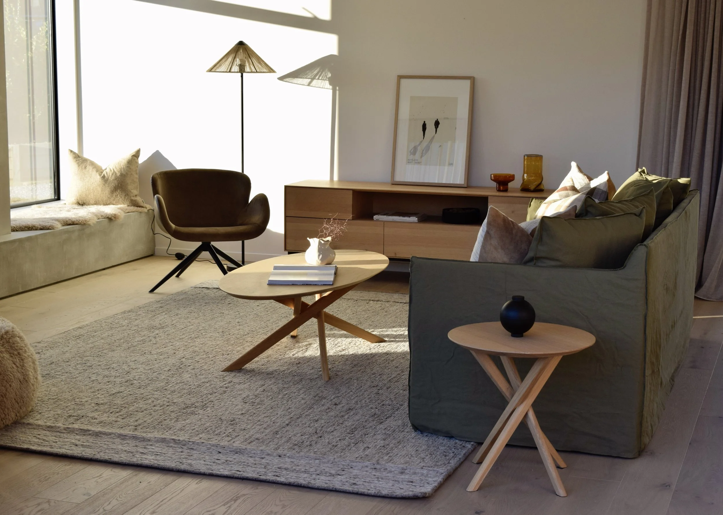 Cozy living room with a window seat, a modern brown armchair, a round wooden coffee table with books, a gray sofa with pillows, a wooden media console with decorative items, and a framed artwork on the wall. Natural light fills the space.
