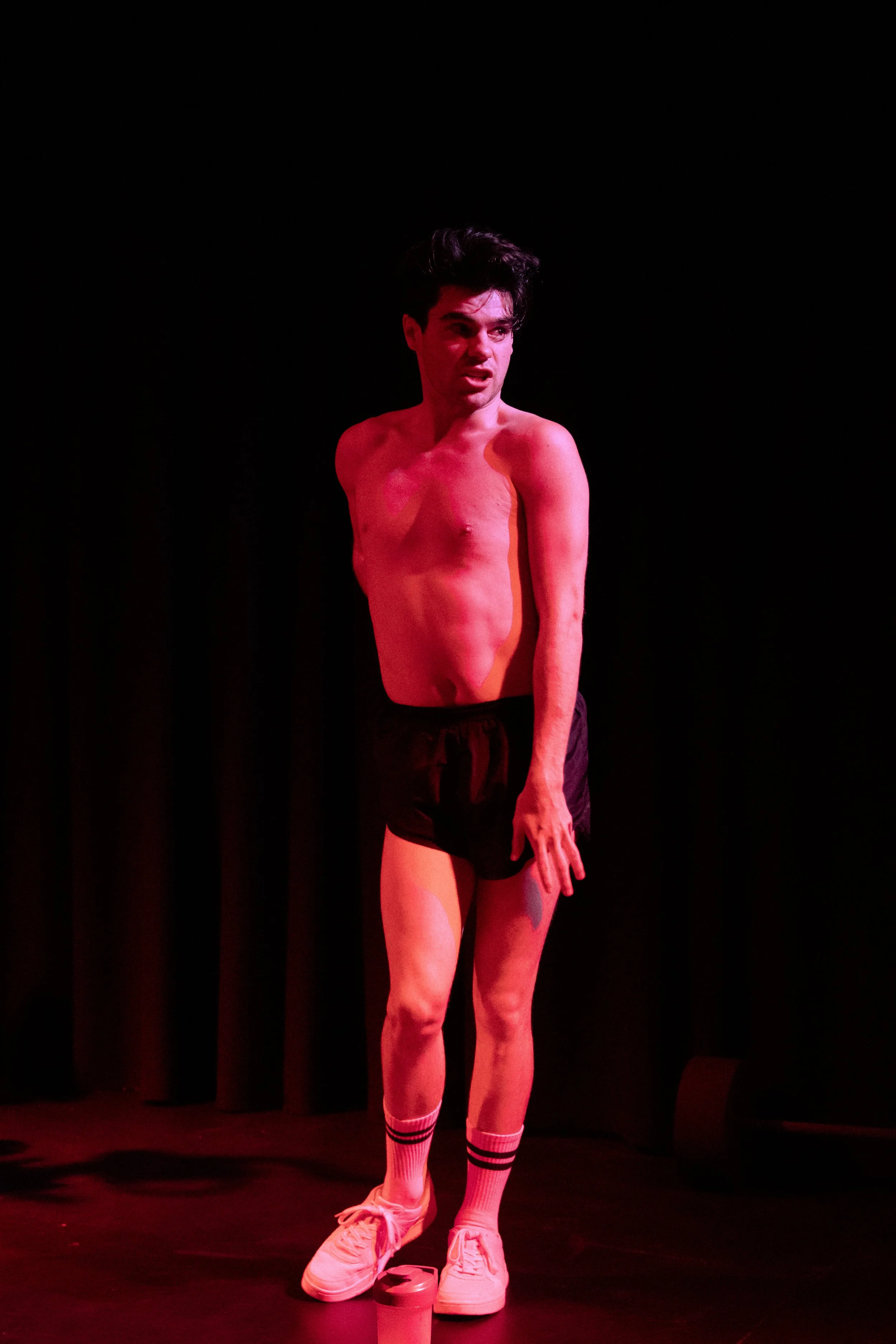 A young man is on stage, shirtless, wearing black shorts, white sneakers, and socks, with a cup on the floor in front of him. He has dark hair and is making a facial expression as if speaking or singing, with a dark background behind him.