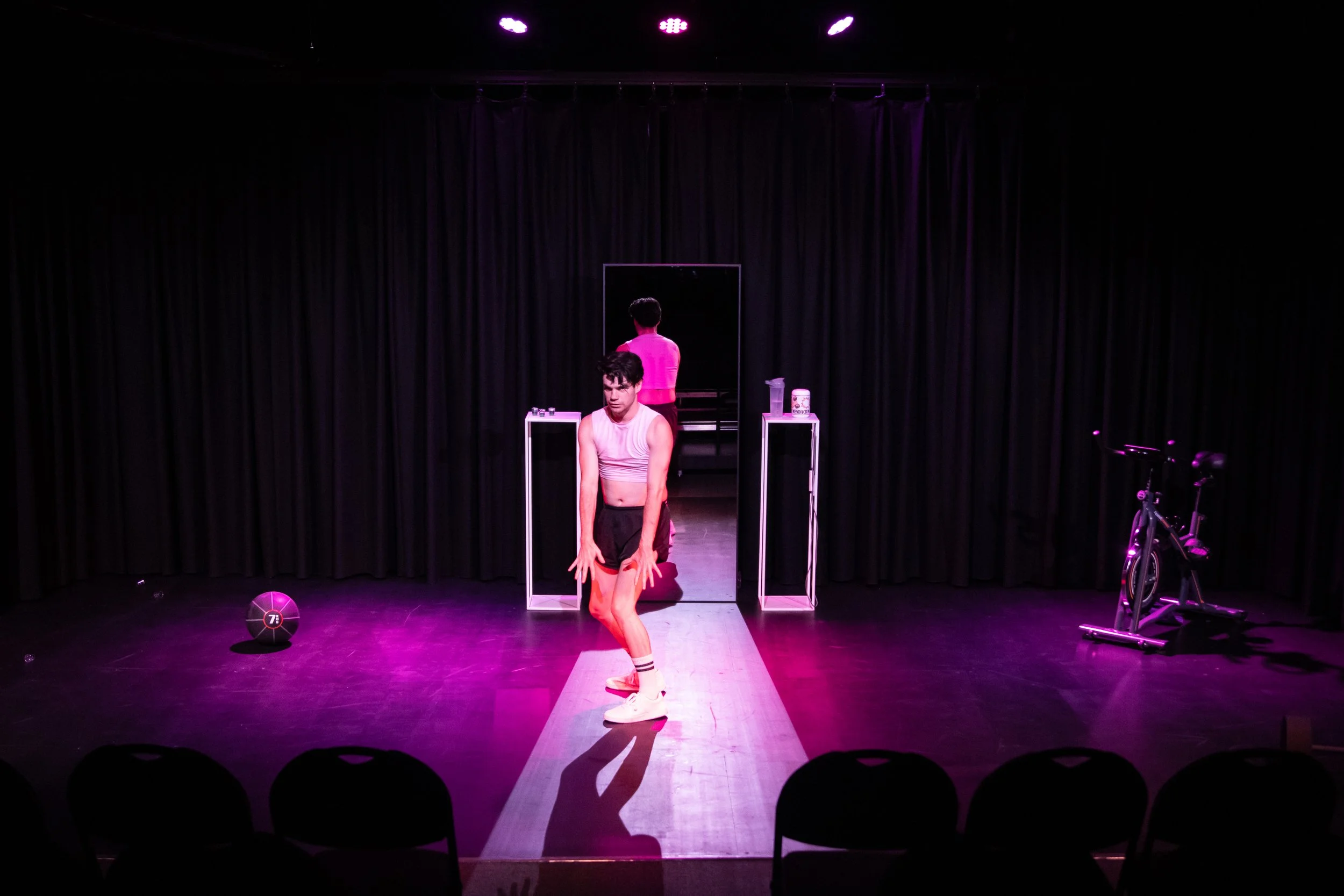 A performer in workout clothing poses on a stage illuminated with pink and purple lighting, with a mirror and props behind him, and a stationary bike off to the right.