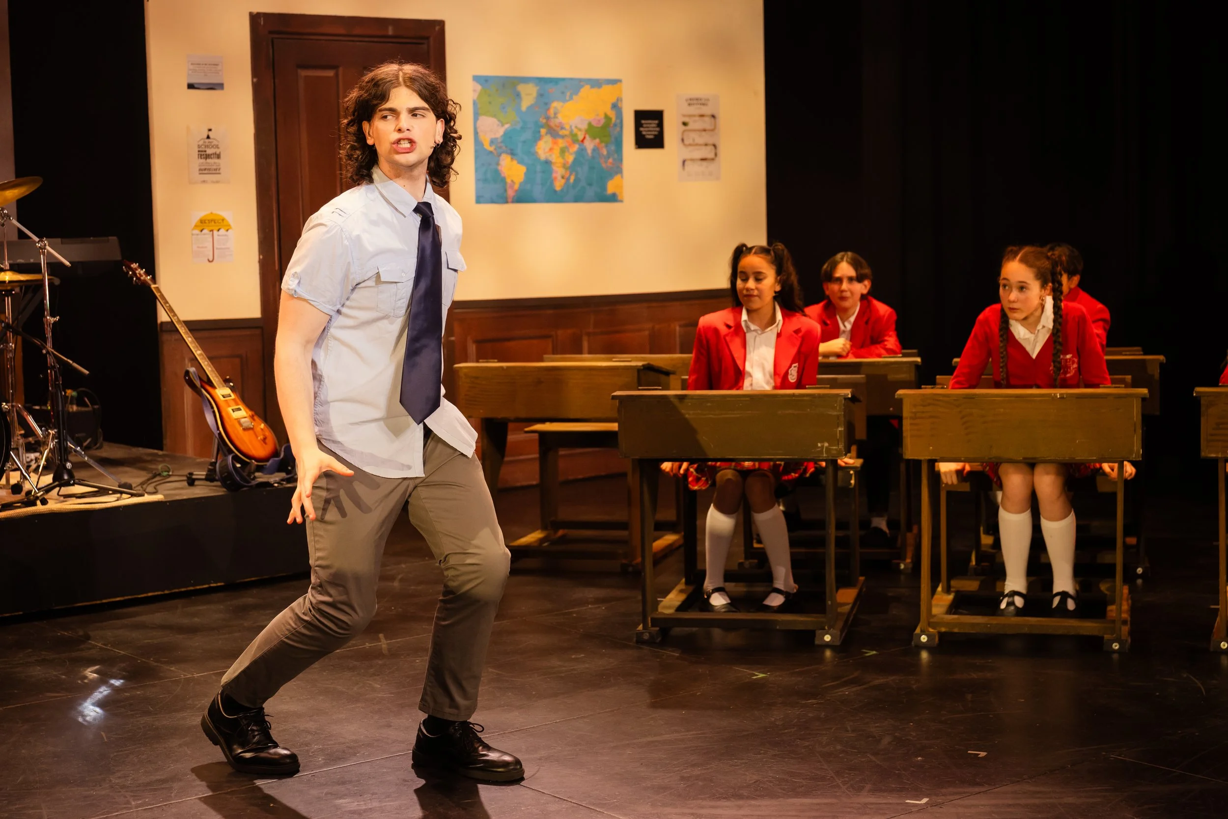 A young man dressed as a student on stage performing in a school play, with children in red uniforms sitting at desks in the background.
