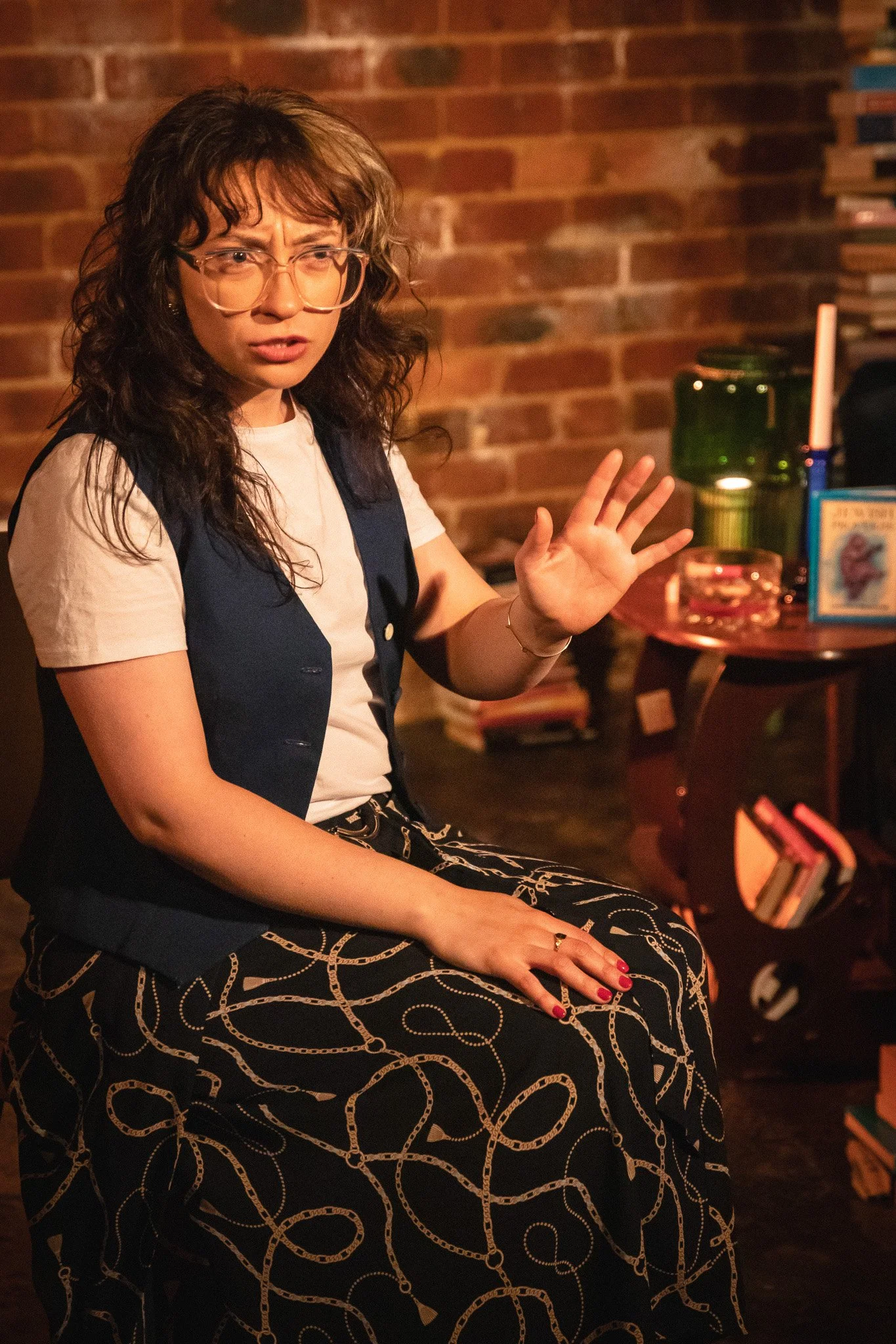 A woman with curly brown hair and glasses sitting on a chair, gesturing with her right hand, in a cozy indoor setting with a brick wall background and a small table with books and decorative items.