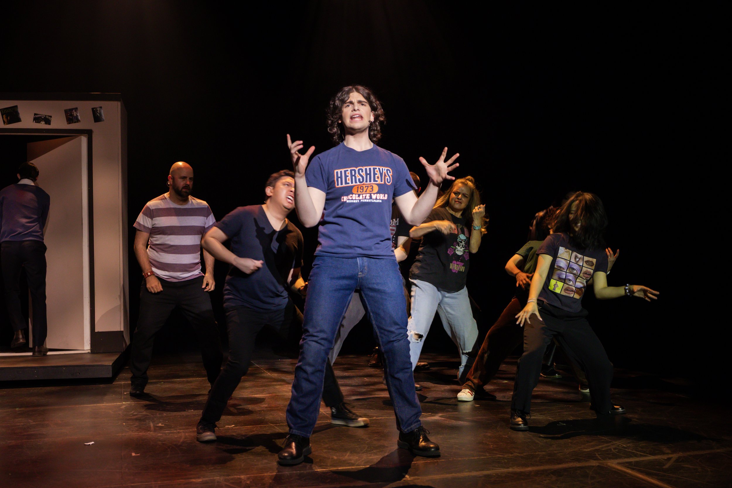 Group of people performing on stage in a theater, with one person in the center wearing a blue Hershey's T-shirt, appearing to sing or speak passionately.