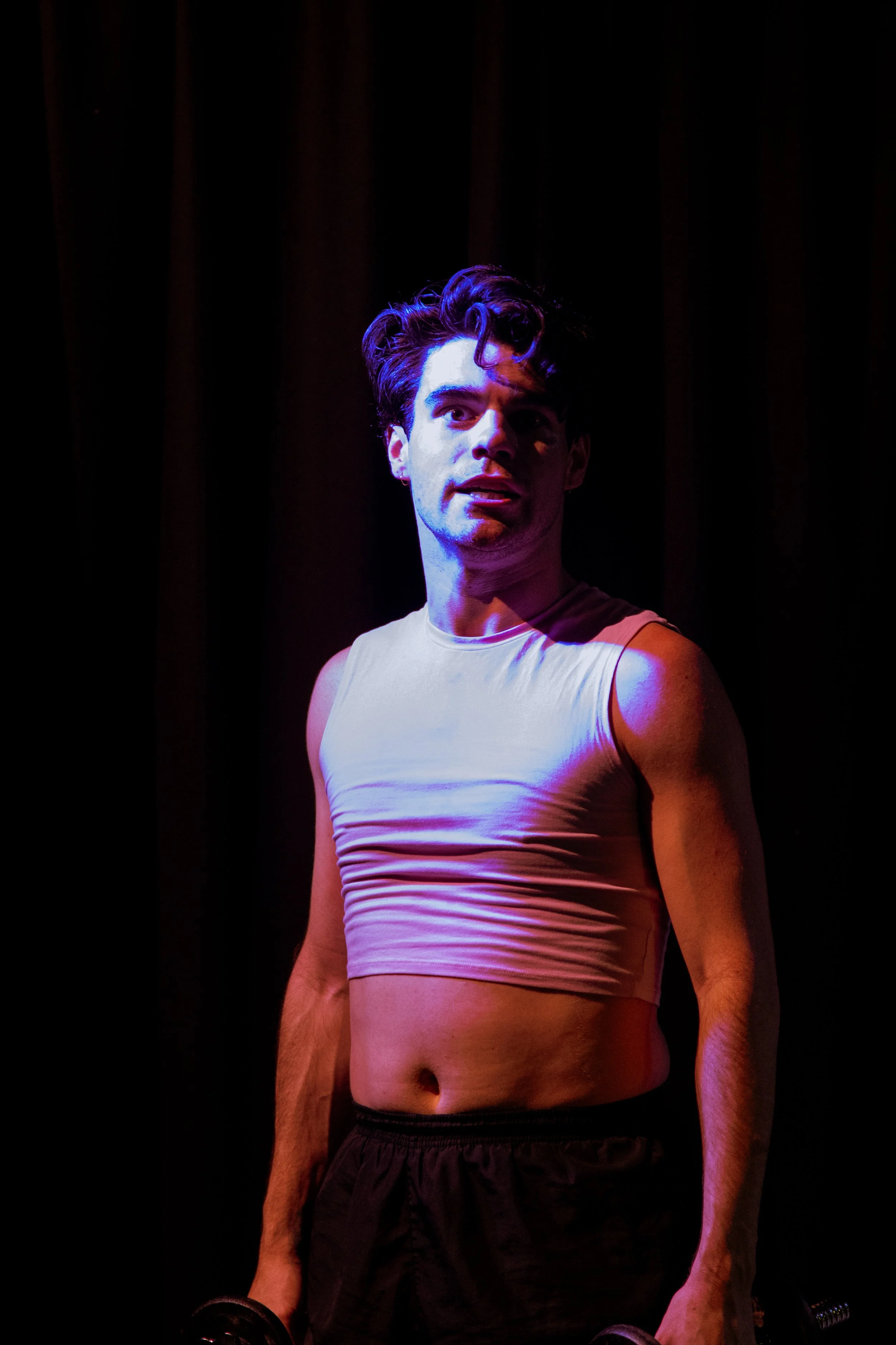 A young man with dark, curly hair wearing a white sleeveless shirt and black shorts, standing with a dark curtain backdrop, illuminated by colorful stage lighting.