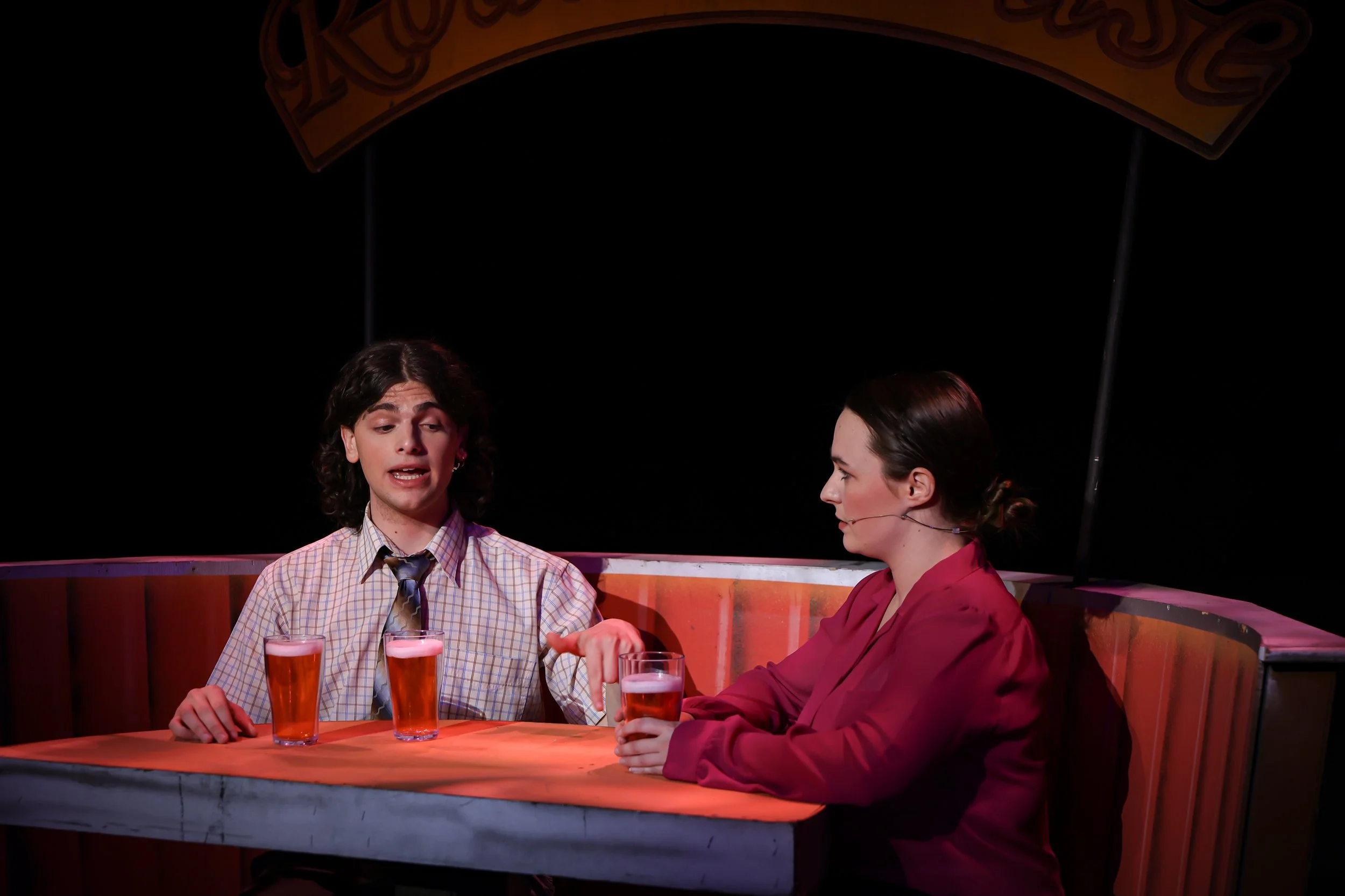Two actors sitting at a table with three glasses of beer, engaging in conversation during a performance on stage.