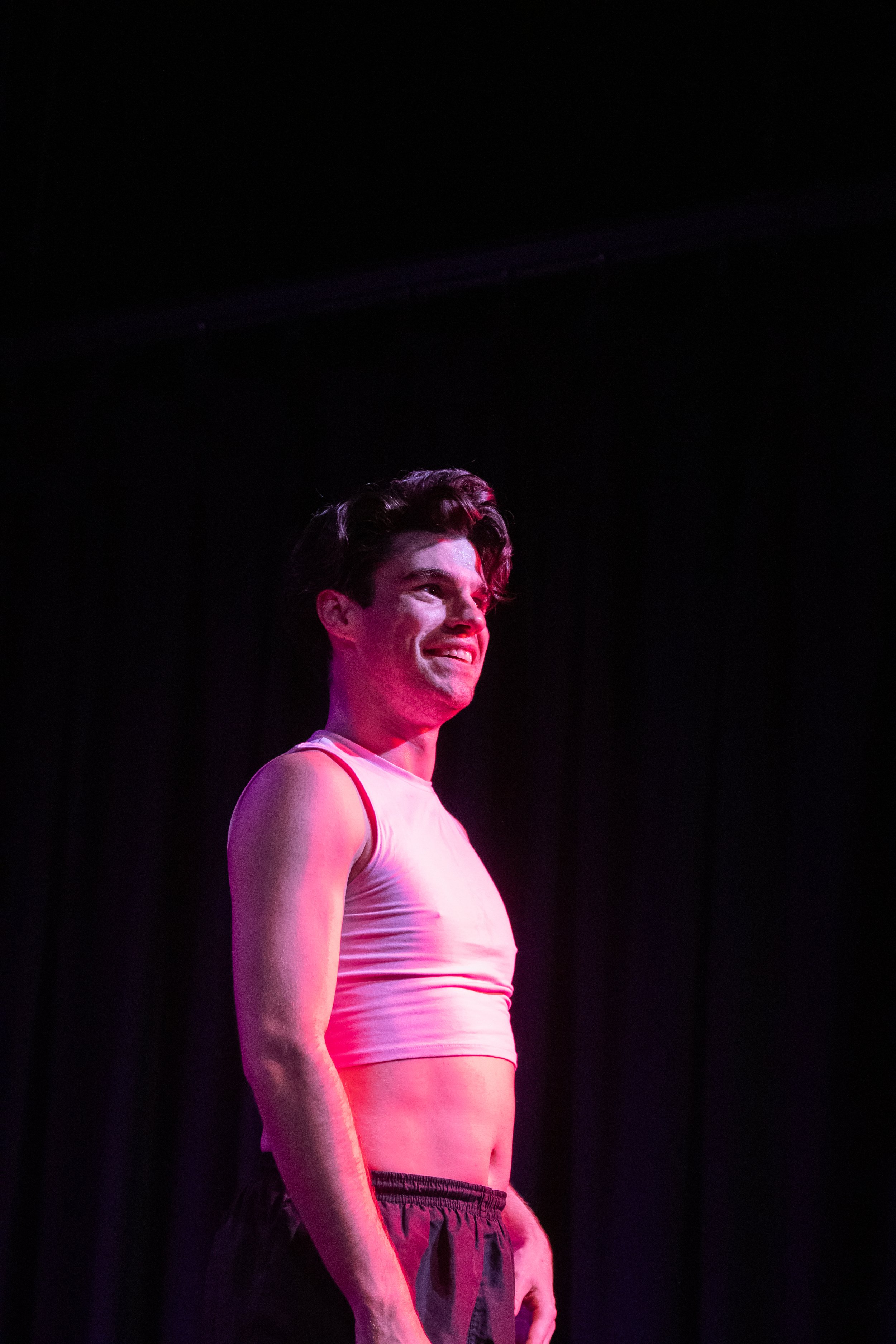 A young man wearing a pink crop top and black shorts standing on a dark stage with pink and purple lighting.