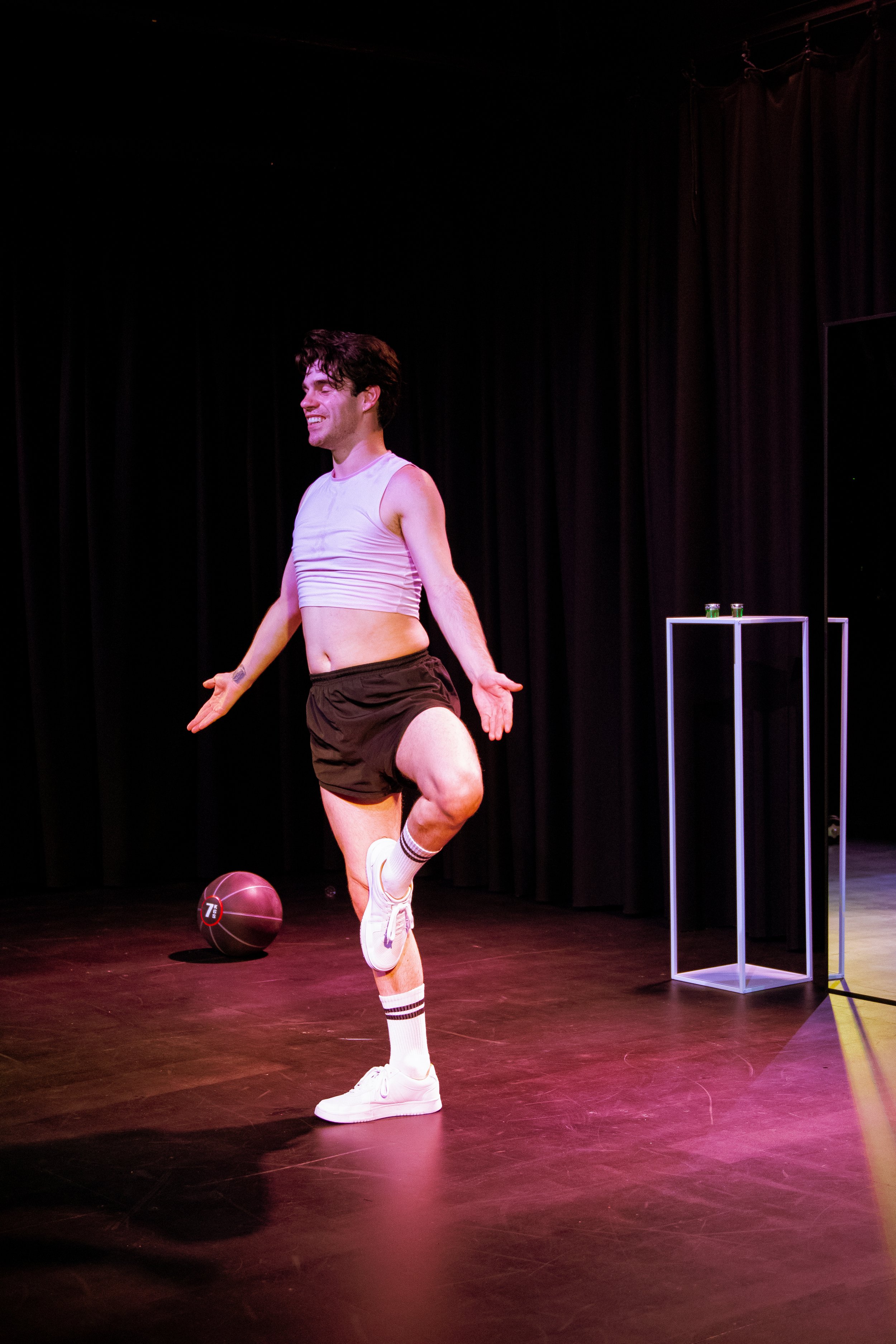 A person standing on stage, wearing a white sleeveless crop top, black shorts, white sneakers, and striped socks, balancing on one leg with a playful expression. There is a medicine ball on the floor and a small table with drinks behind them, with a 