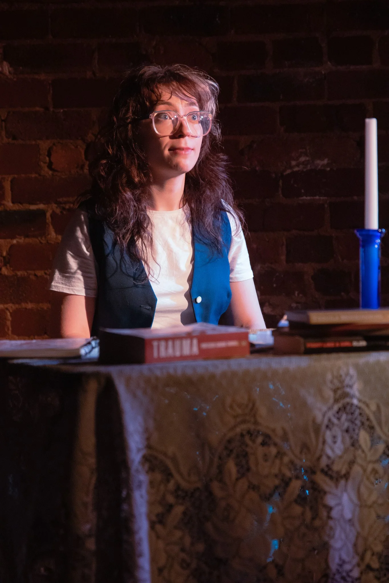 A young woman with glasses and curly hair sitting at a table with books, a candle, and a box labeled "Trauma" in front of her, against a brick wall background.