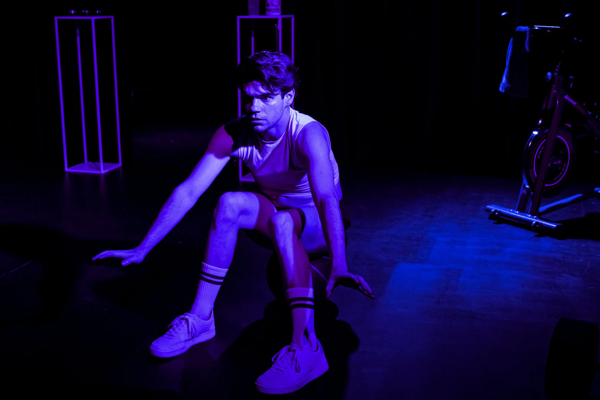 A young man with messy hair, wearing a sleeveless shirt, shorts, and sneakers, is sitting on the floor in a dark room with blue lighting. There are purple geometric structures and a stationary exercise bike in the background.