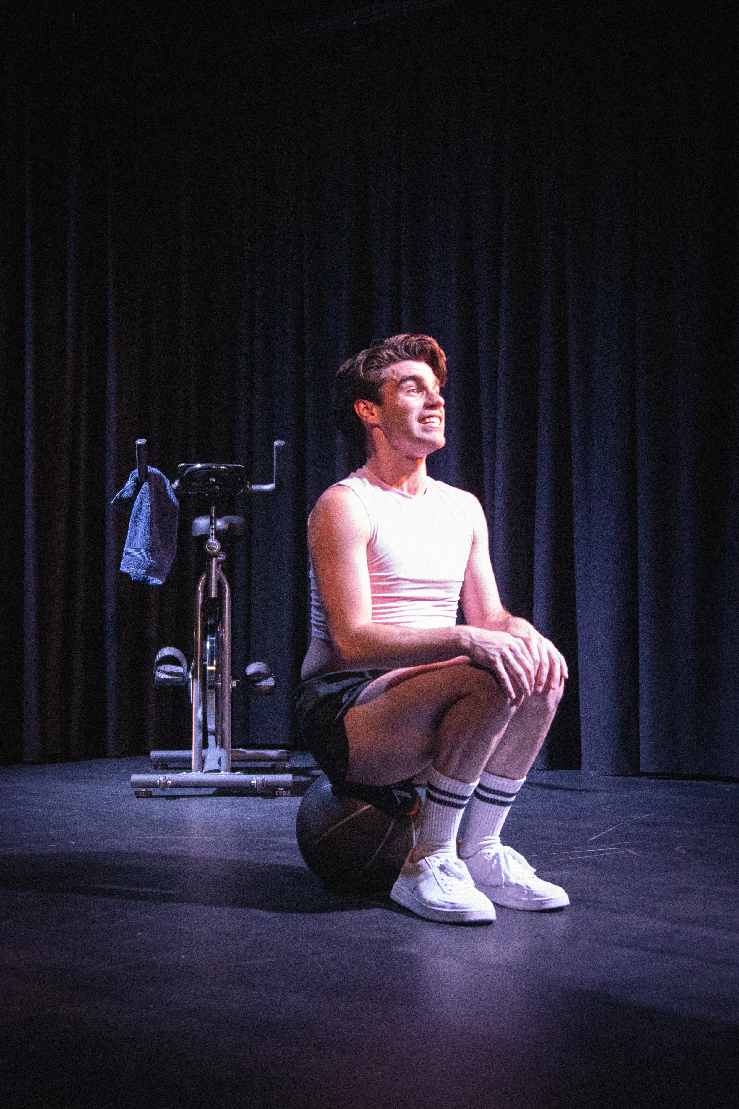 A male performer kneeling on a stage with a black curtain background, wearing a white tank top, black shorts, white socks, and white sneakers, smiling while holding a black medicine ball, with an exercise bike behind him.