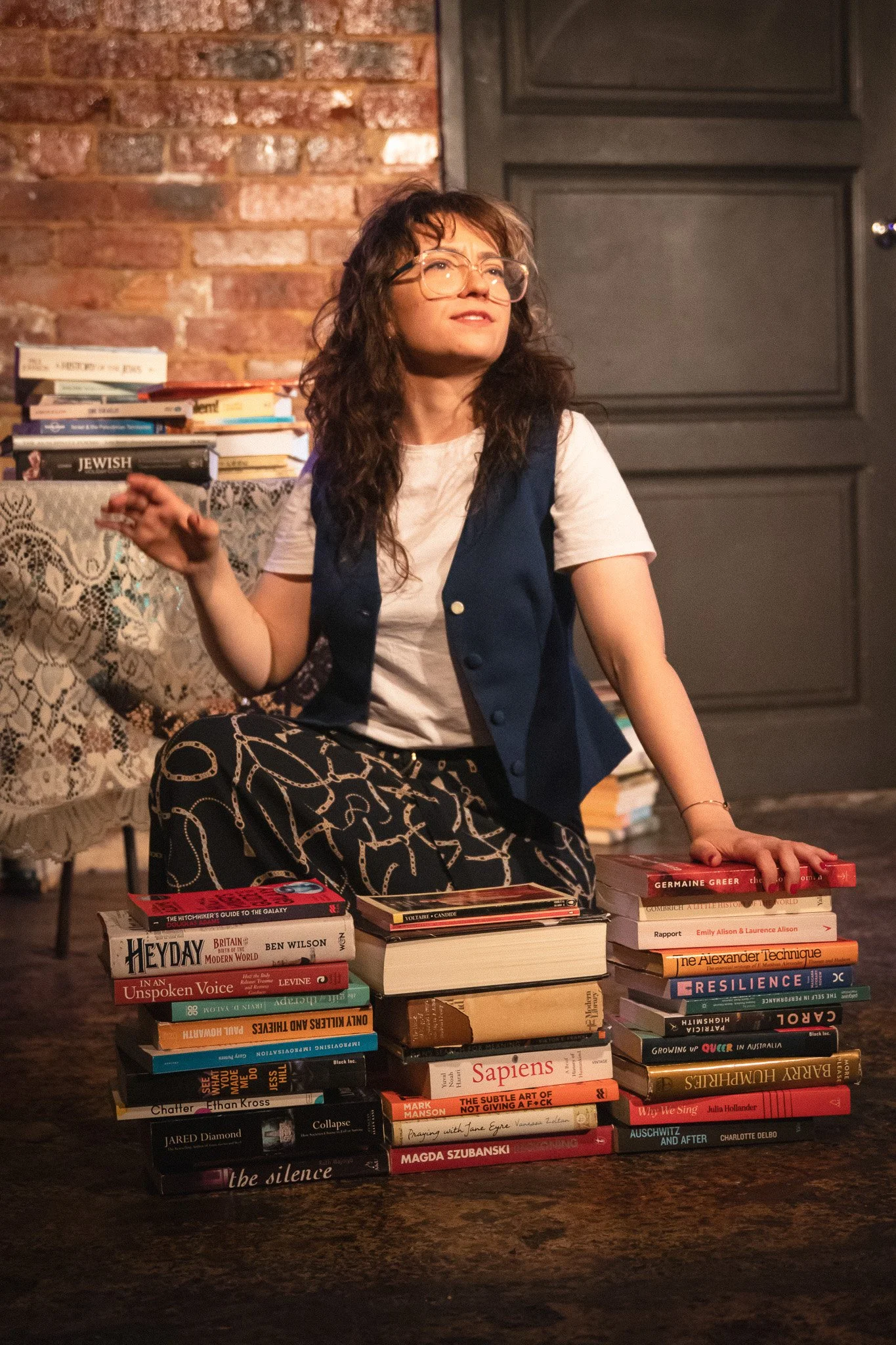 A woman with glasses and wavy hair, sitting on the floor behind two stacks of books, in a room with a brick wall and a dark wooden door.