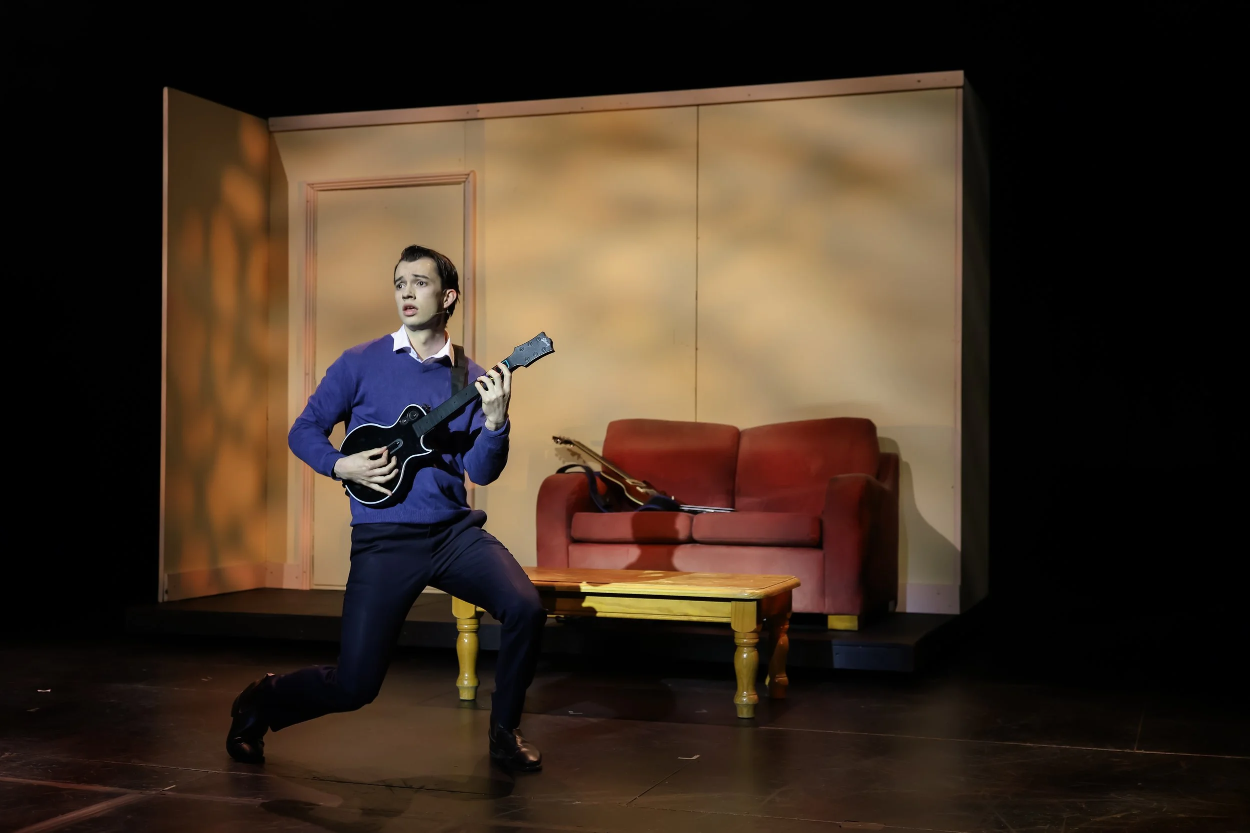 A man performing on stage with a guitar, in front of a set resembling a living room with a red sofa and a wooden coffee table.