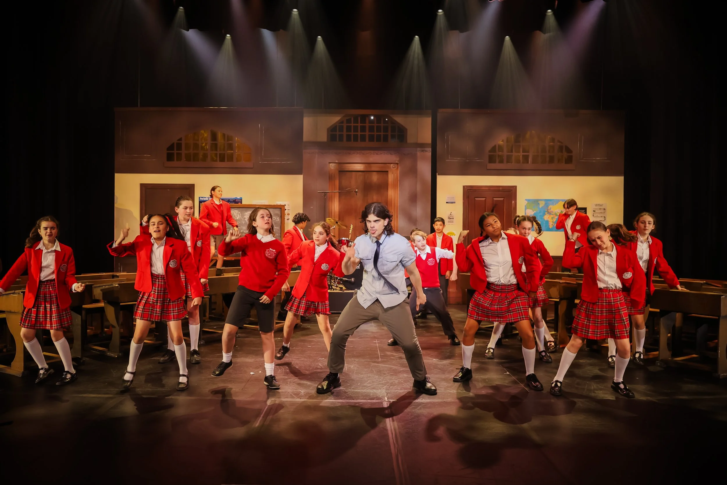 Students in red uniforms perform a dance on stage set as a school classroom, with a male student in casual clothes leading the dance.
