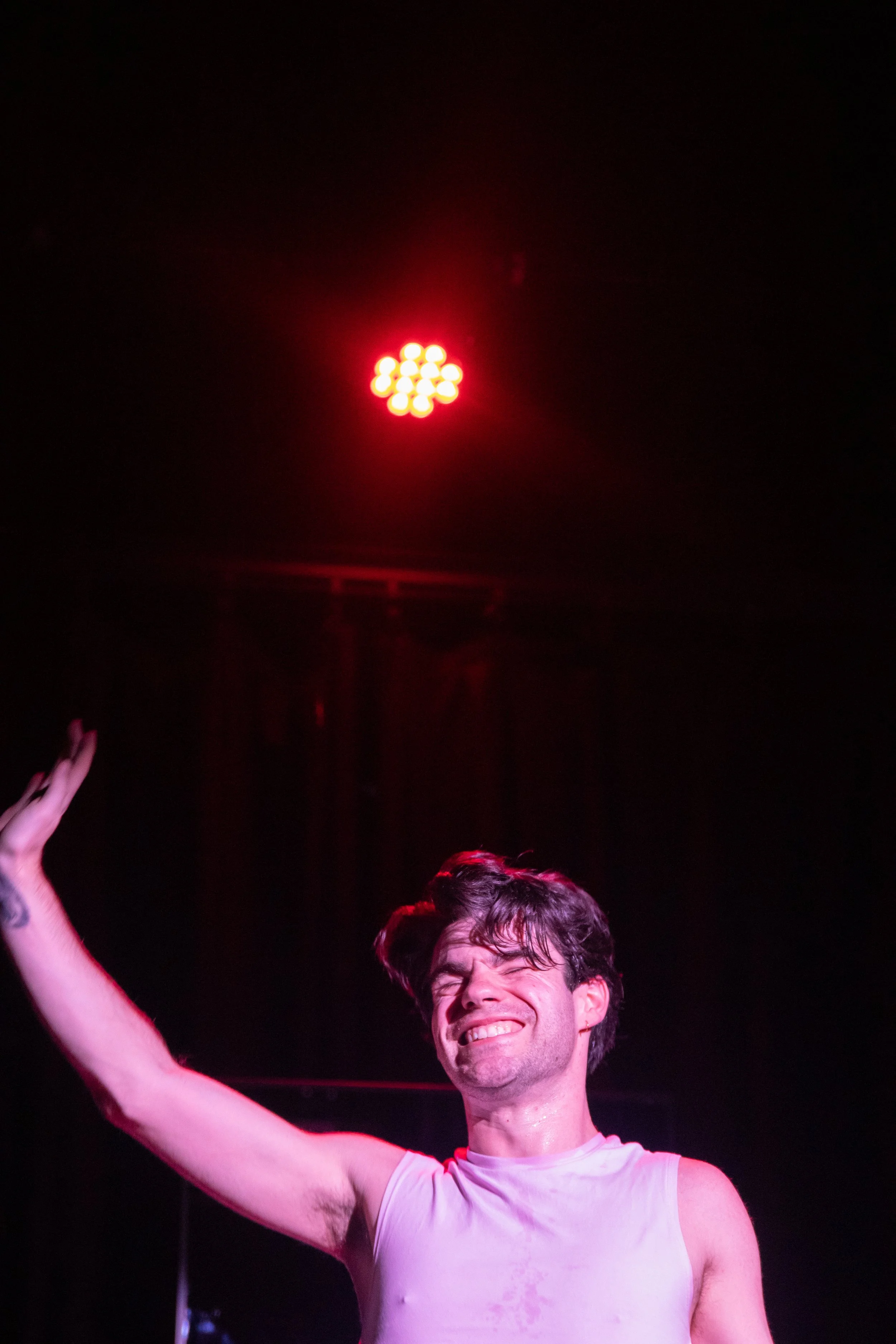 A man in a white tank top smiling and waving with a red stage light overhead.