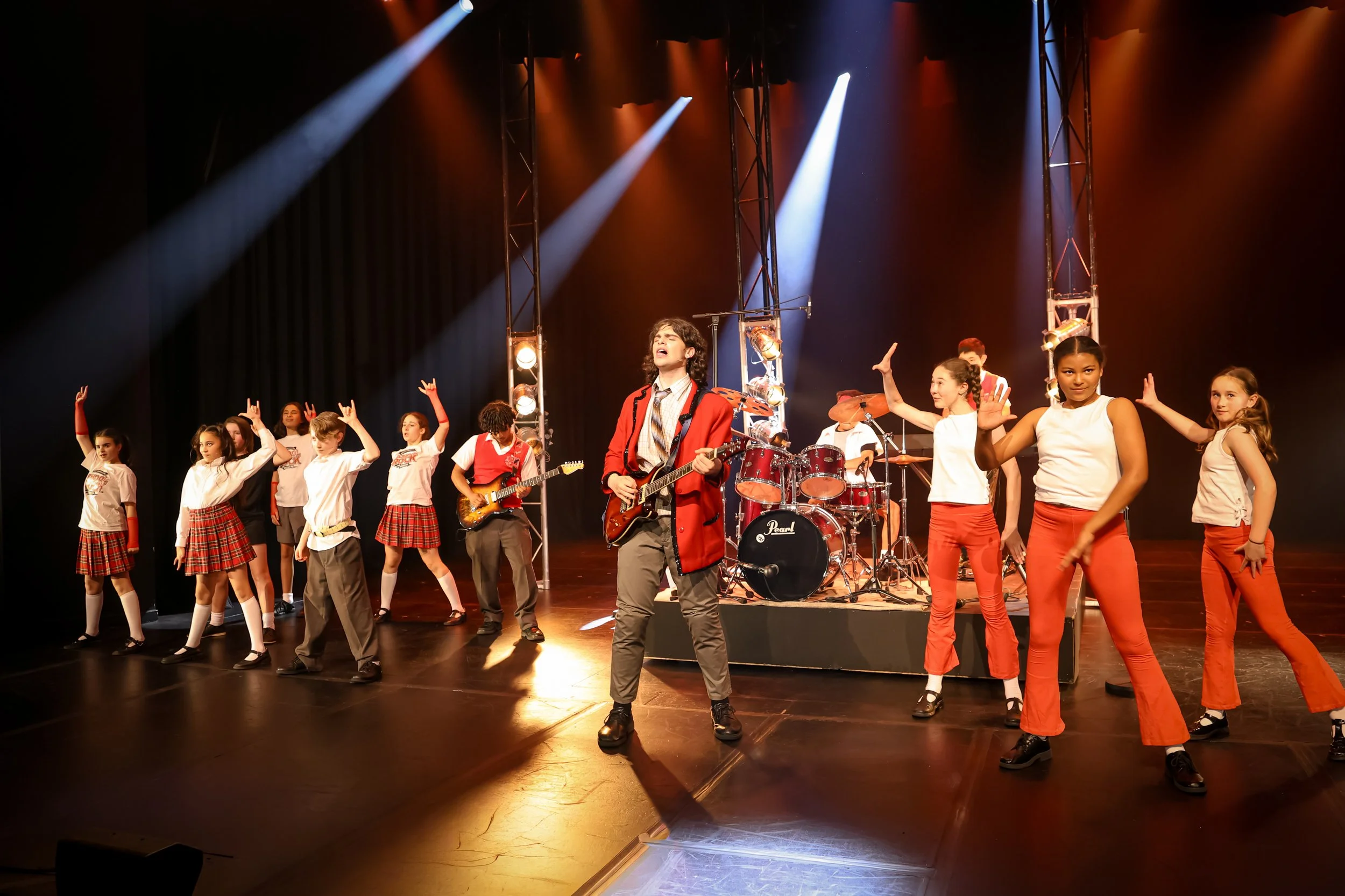 Group of children and teenagers performing on stage with musical instruments, under colorful stage lighting, with a girl in the center singing and playing guitar.