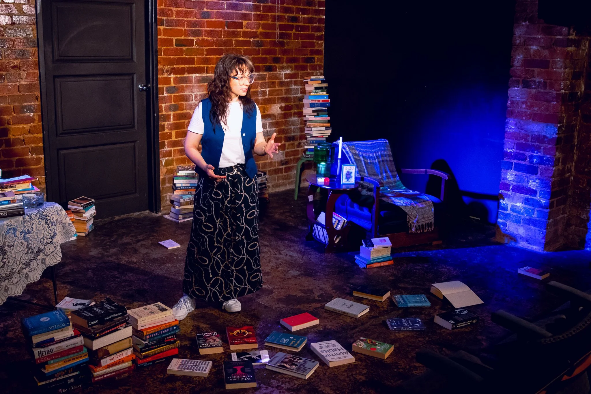 A woman with glasses stands in a room with brick walls, surrounded by scattered books and stacked books, some on tables and others on the floor. She is wearing a white t-shirt under a blue vest and patterned black and white pants. The room has a cozy