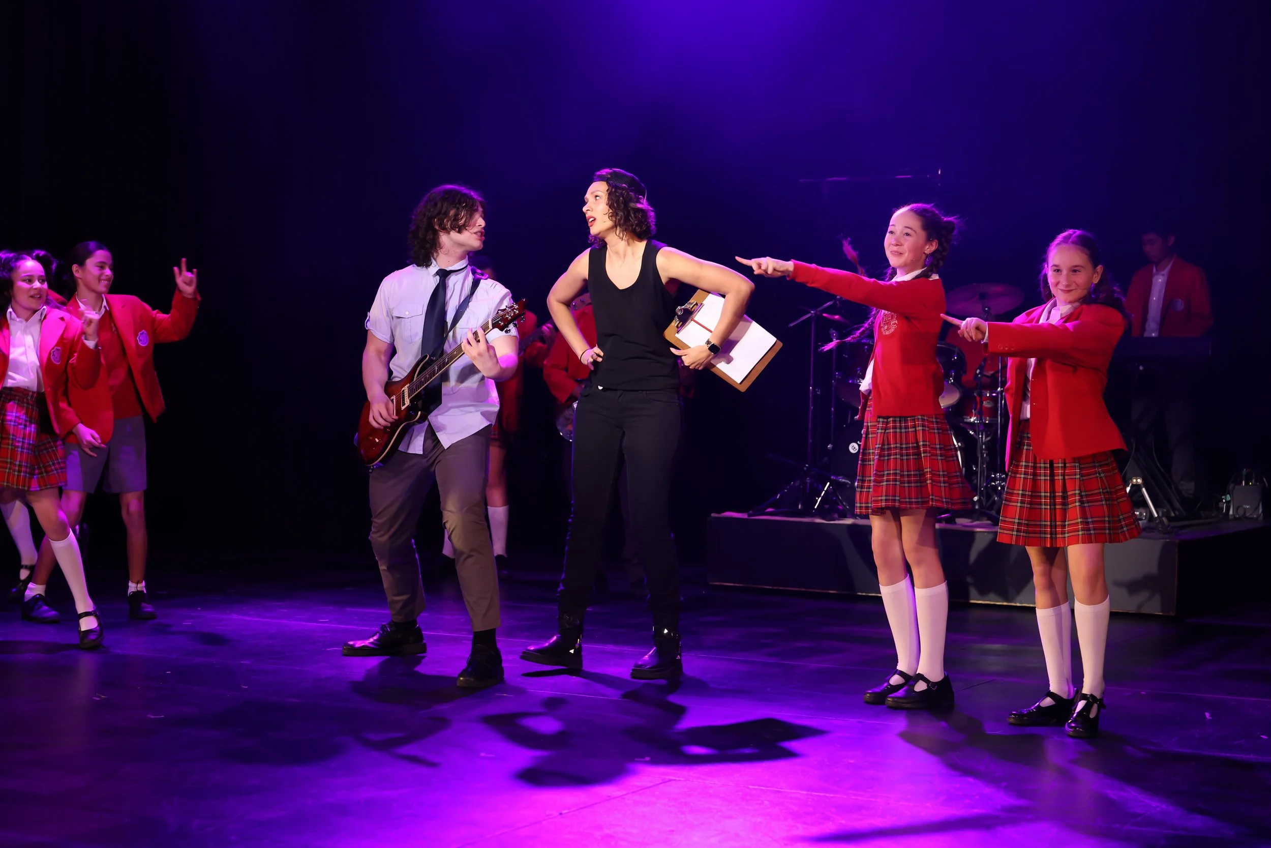 Scene from a school talent show or musical on stage with children and young adults performing, some wearing red plaid skirts and school uniforms, others with musical instruments, under purple stage lighting.