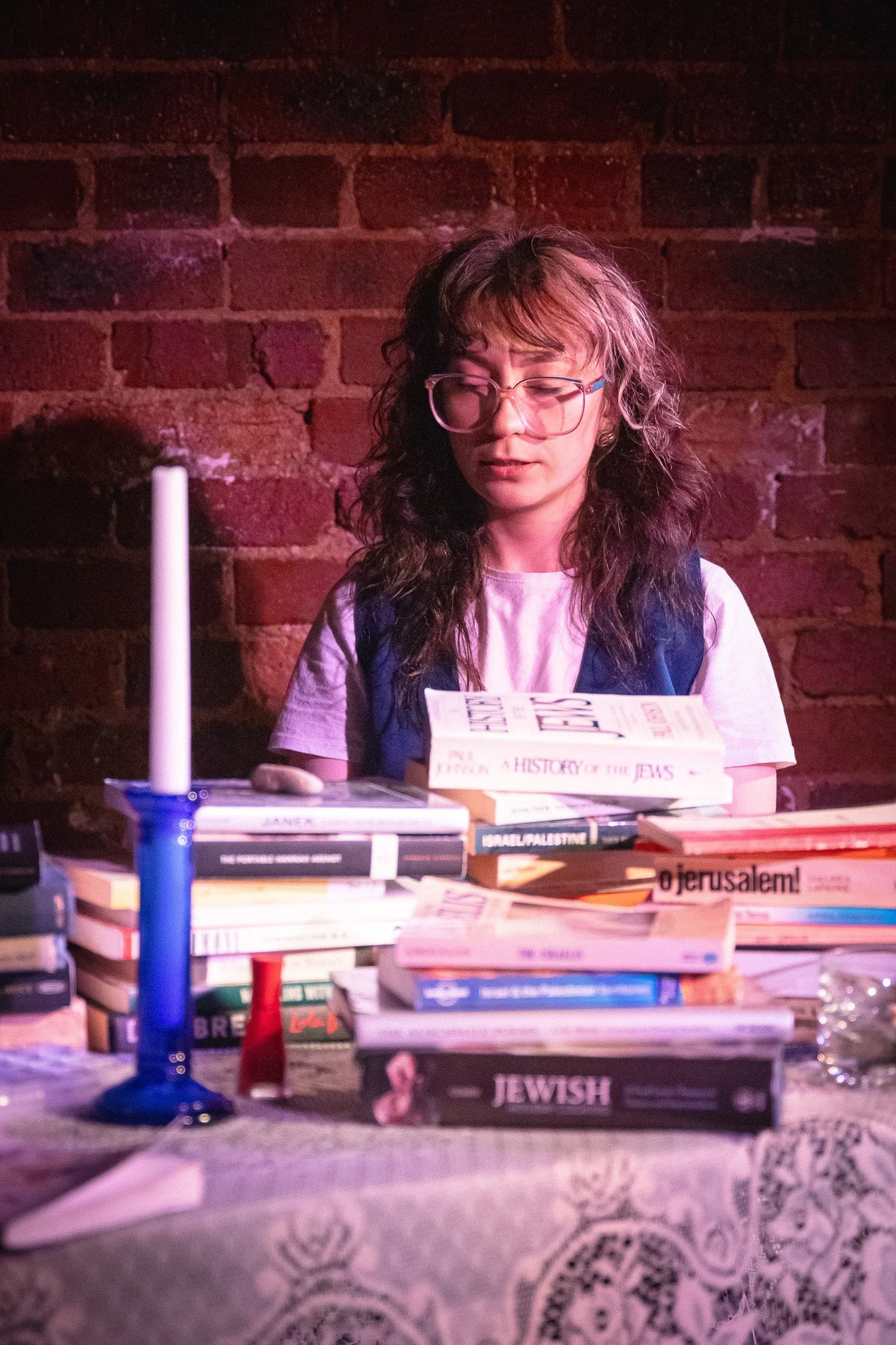 A young woman with curly hair and glasses reading a book at a table filled with many books, with a brick wall background.