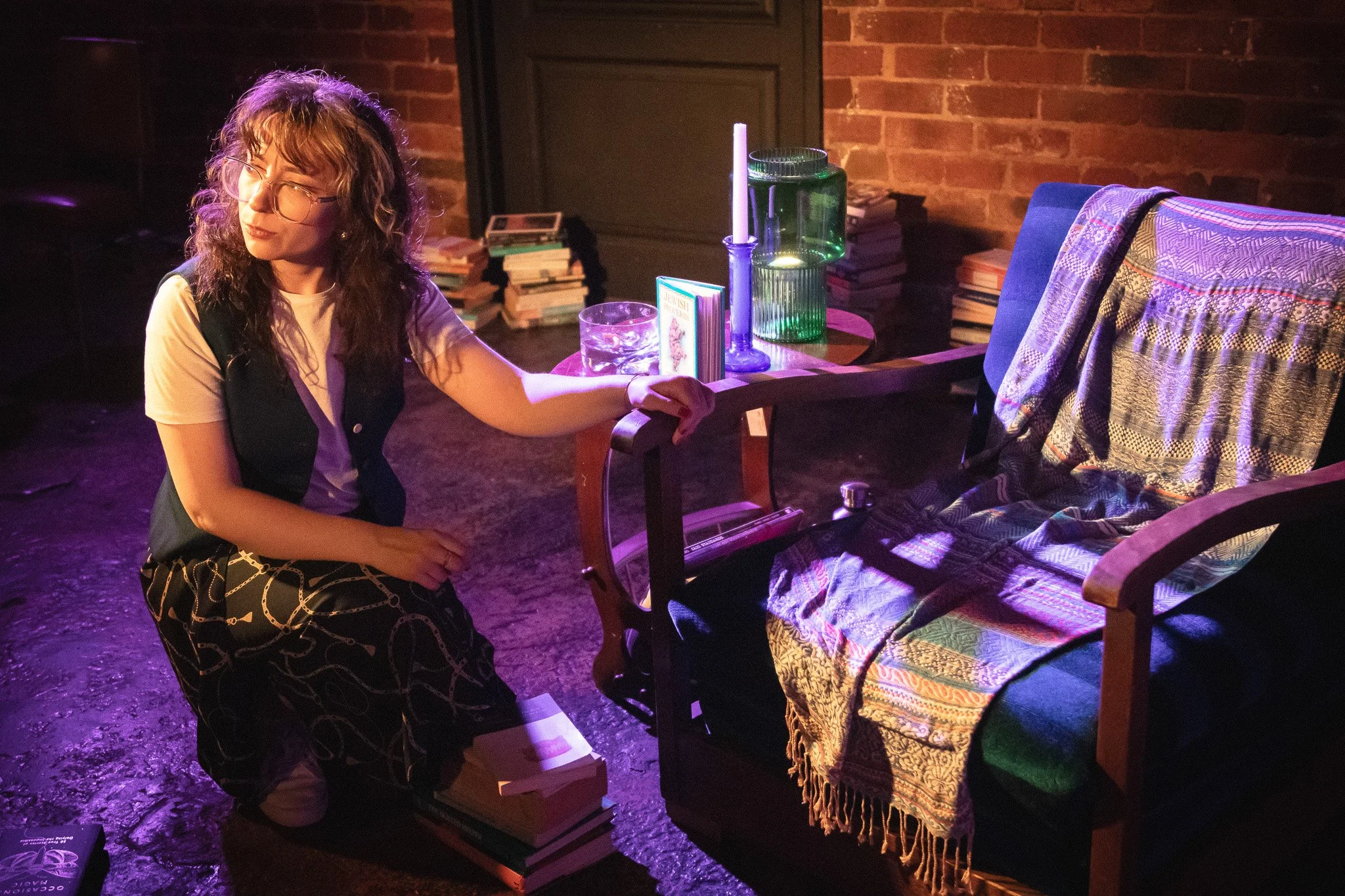 A woman with curly hair and glasses kneels next to a stack of books in a dimly lit room with a brick wall. She is holding the arm of a wooden chair draped with a patterned fabric. There are more books and decorative items like candles and glassware o