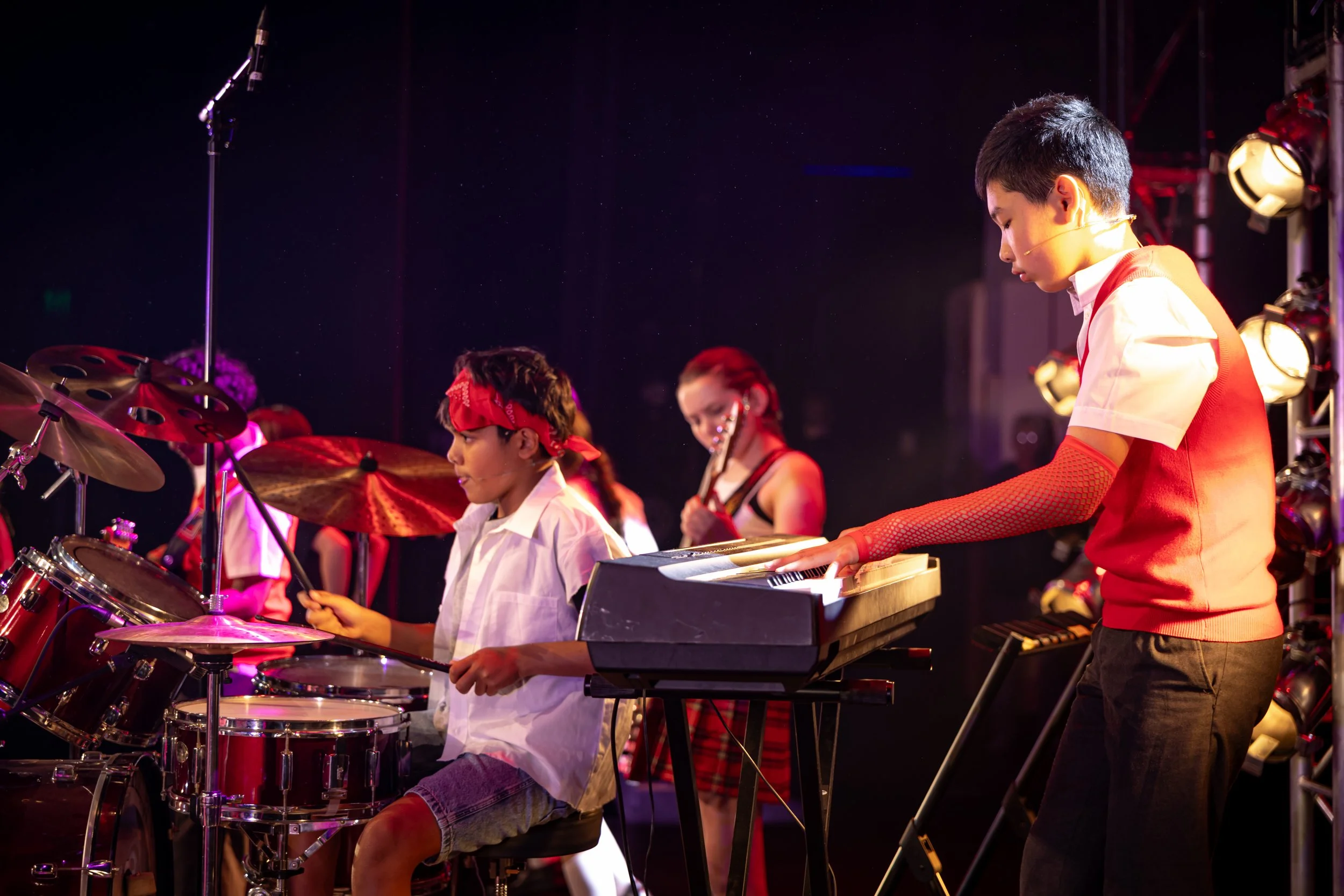 Children performing music on stage, playing drums and keyboard, with a guitarist in the background, under stage lights.