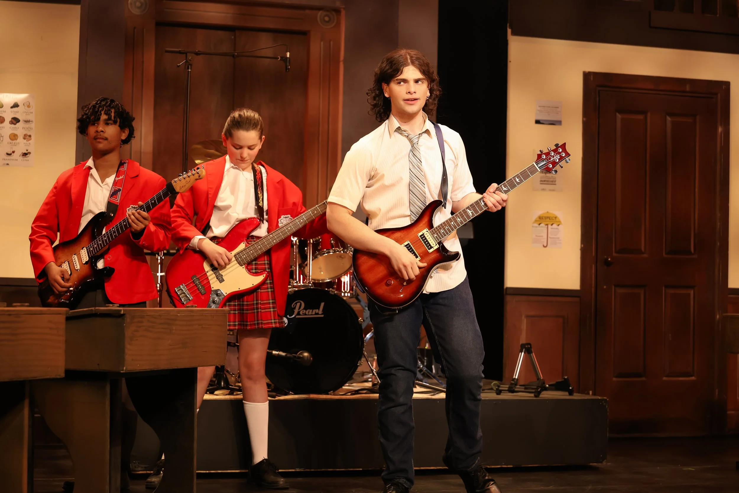 Four teenagers perform with guitars on a stage in a music classroom or rehearsal space, with a drum set in the background, wooden walls, and closed doors.