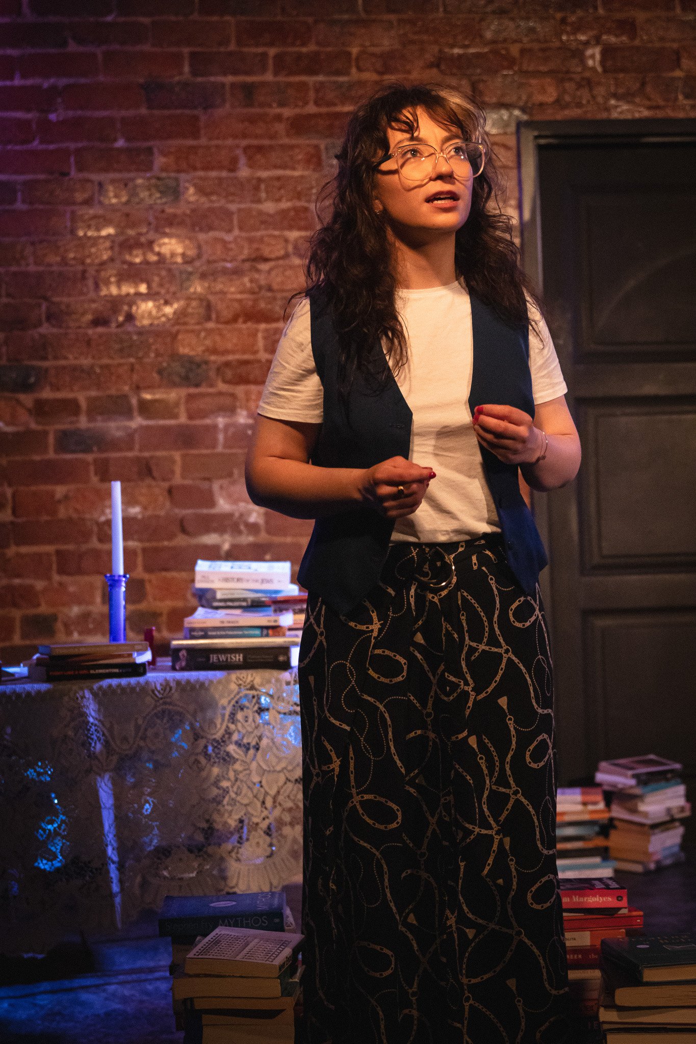 A woman with glasses and curly dark hair, wearing a white t-shirt and patterned pants, stands in front of a brick wall, appearing to speak or perform. There are stacks of books and a lit candle on a table behind her.