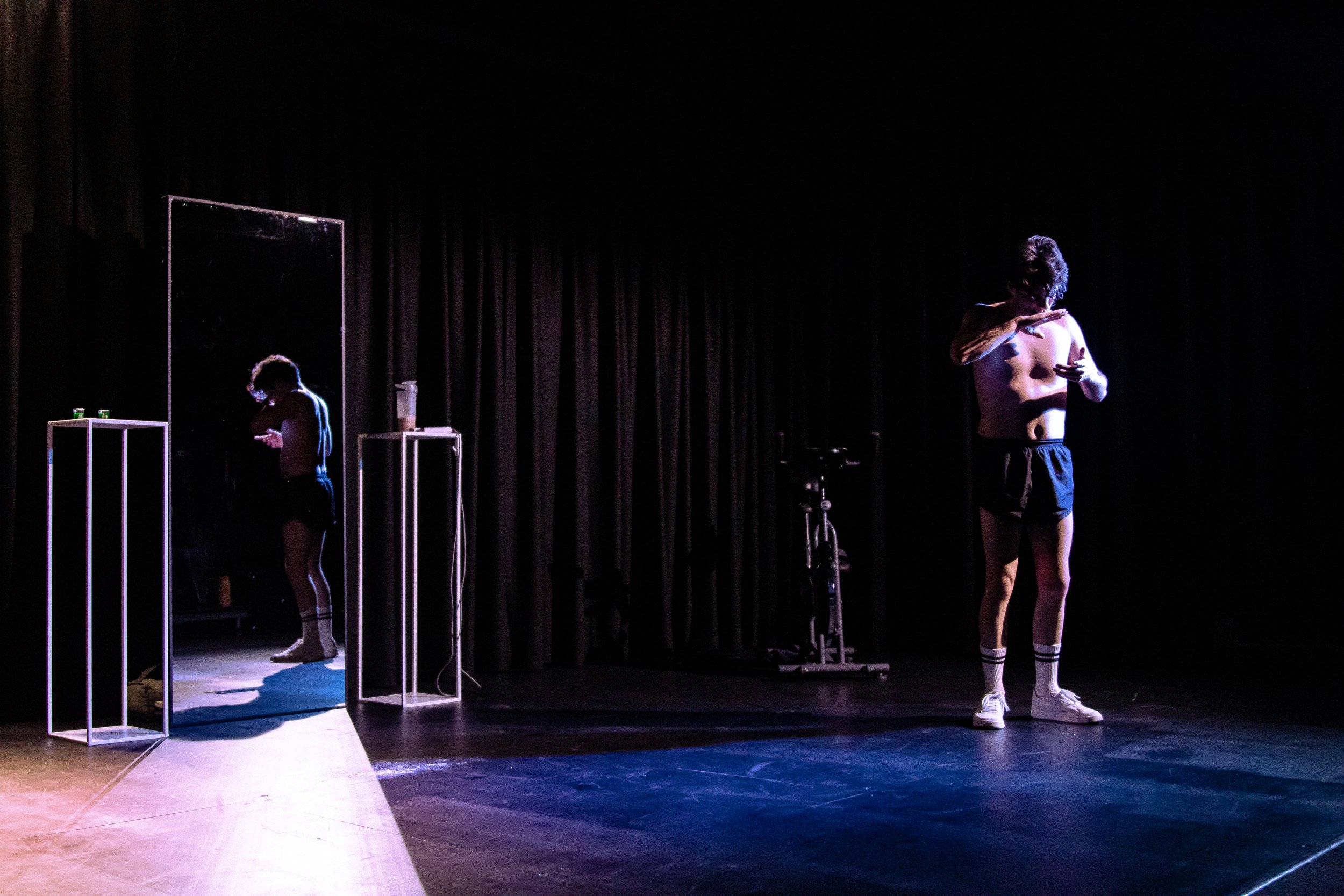 A shirtless man wearing shorts, socks, and sneakers standing on a stage with black curtains in the background. He is looking down and appears to be adjusting his hoodie. A mirror and two small tables with objects on top are on the left side of the st
