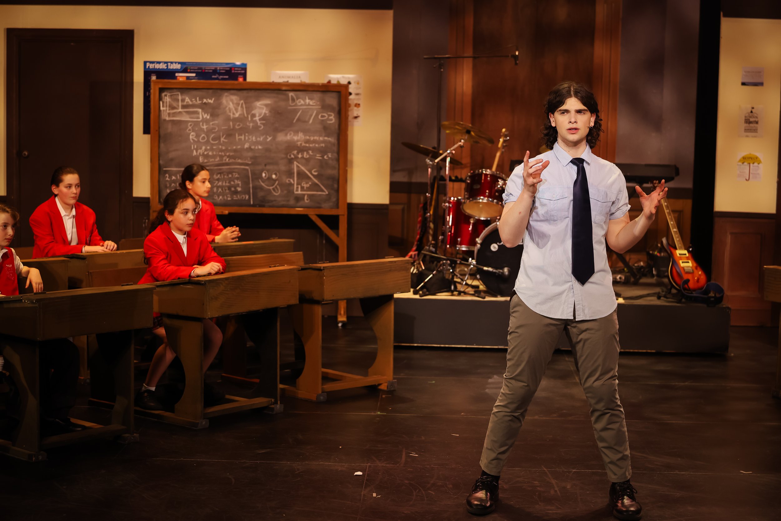 A young man in a schoolboy uniform with a tie is speaking or performing in front of a classroom with a blackboard, musical instruments, and students in red uniforms seated at desks.