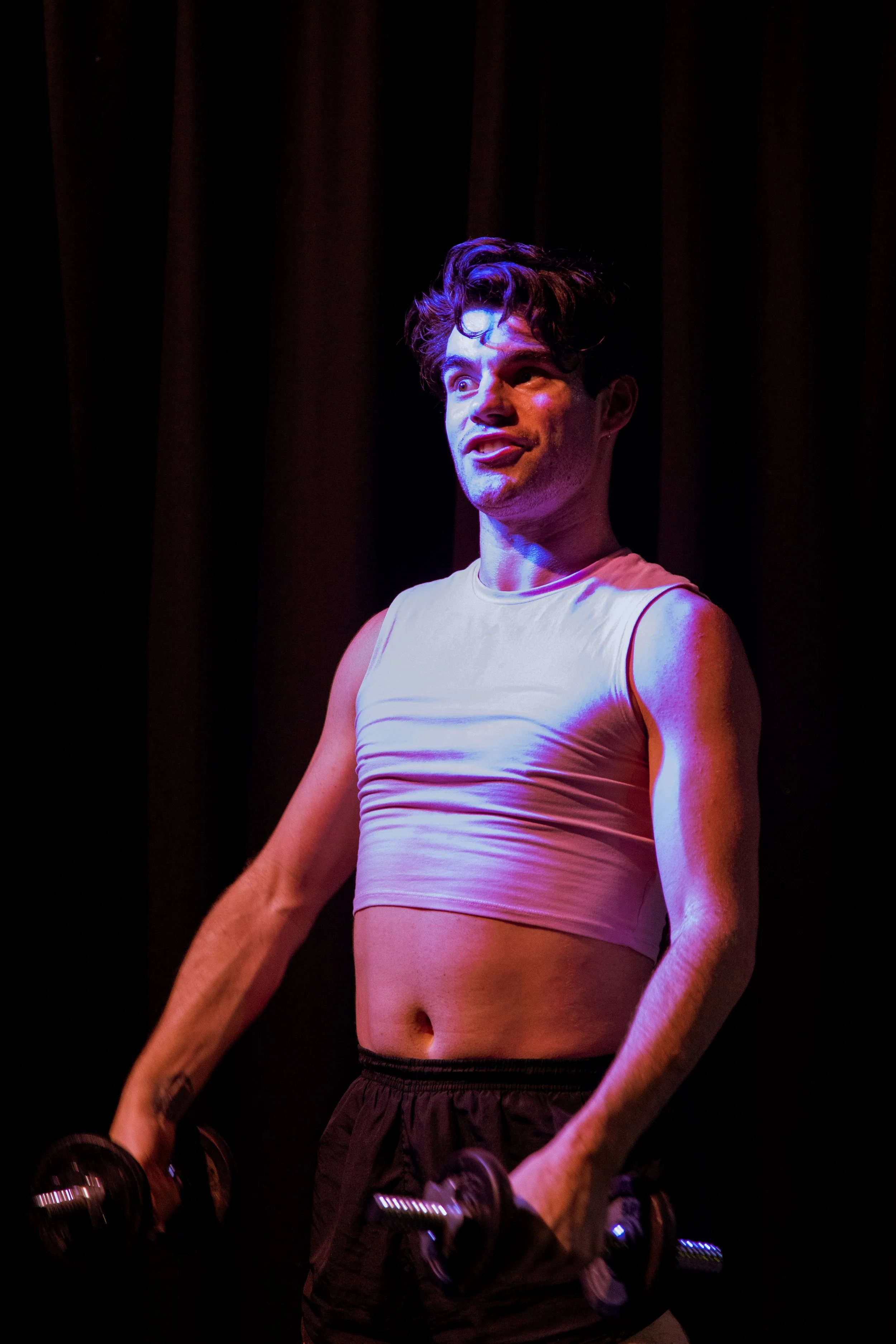 A man with styled dark hair holding dumbbells, wearing a sleeveless white crop top and dark shorts, standing on stage with a dark curtain backdrop.