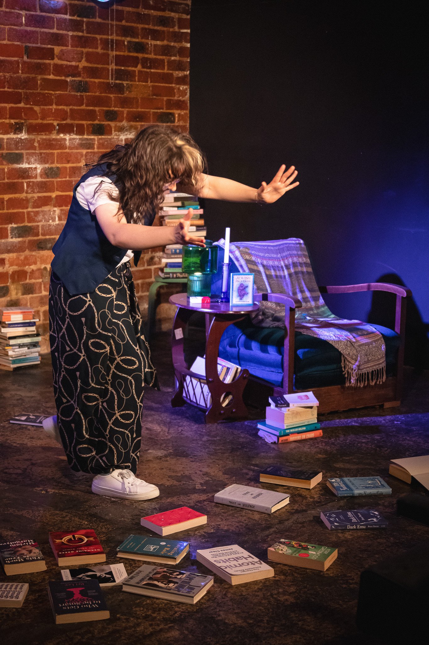 A woman with curly hair, wearing a vest and patterned pants, is standing on a stage surrounded by books on the floor. She appears to be in a theatrical or storytelling performance, with a brick wall behind her and a chair covered with a blanket in th