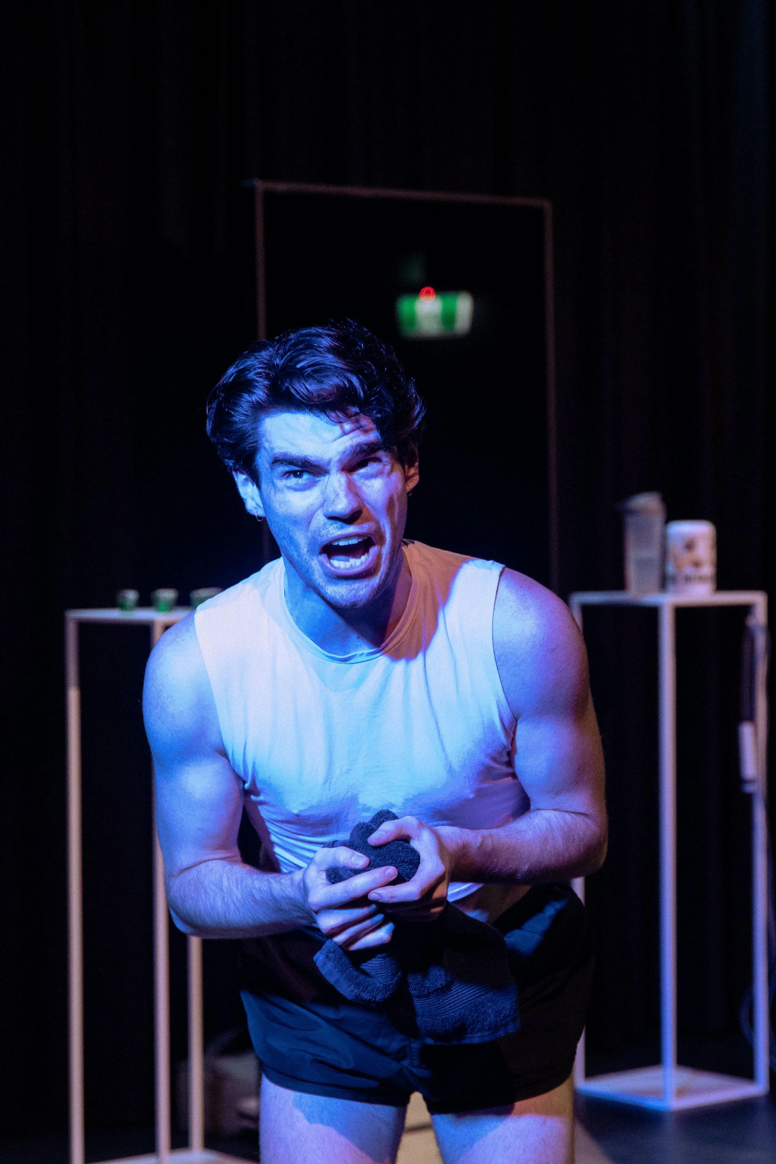 A man with dark hair wearing a white tank top and black shorts, making an intense expression on stage with a dark background and minimal props.