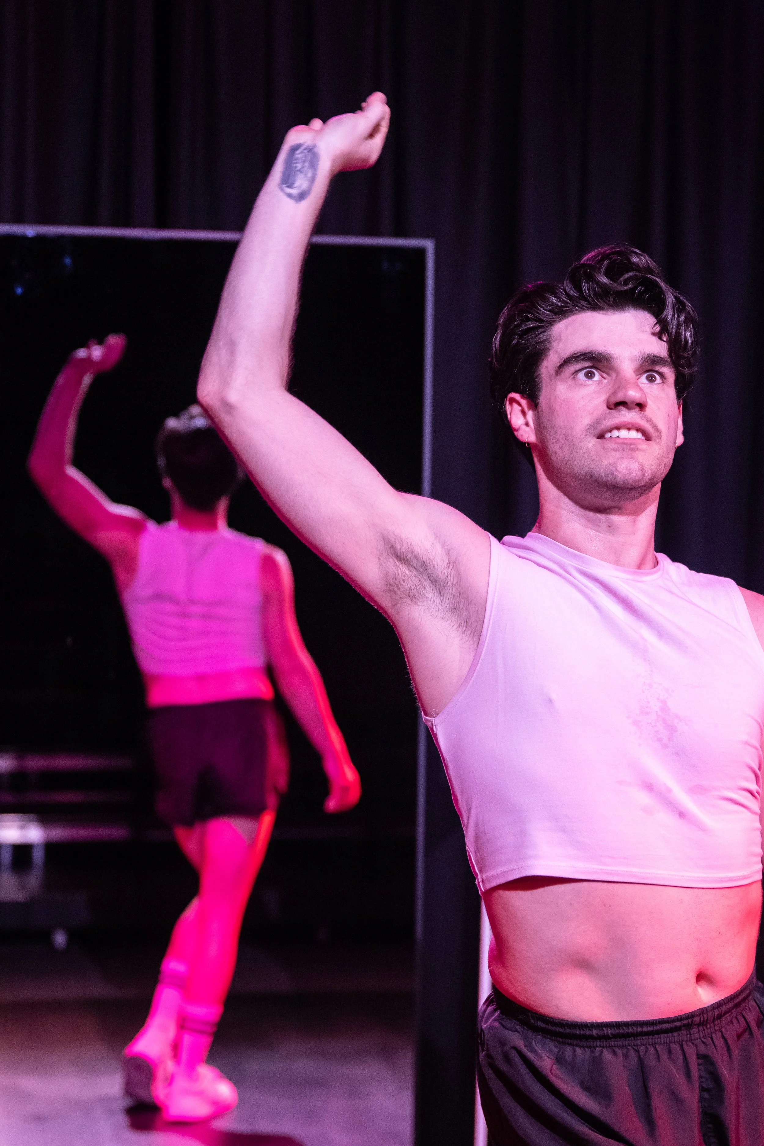 A young man looking at his reflection in a mirror, flexing his arm with a serious expression. In the background, a young woman faces the mirror, stretching her arm upward.