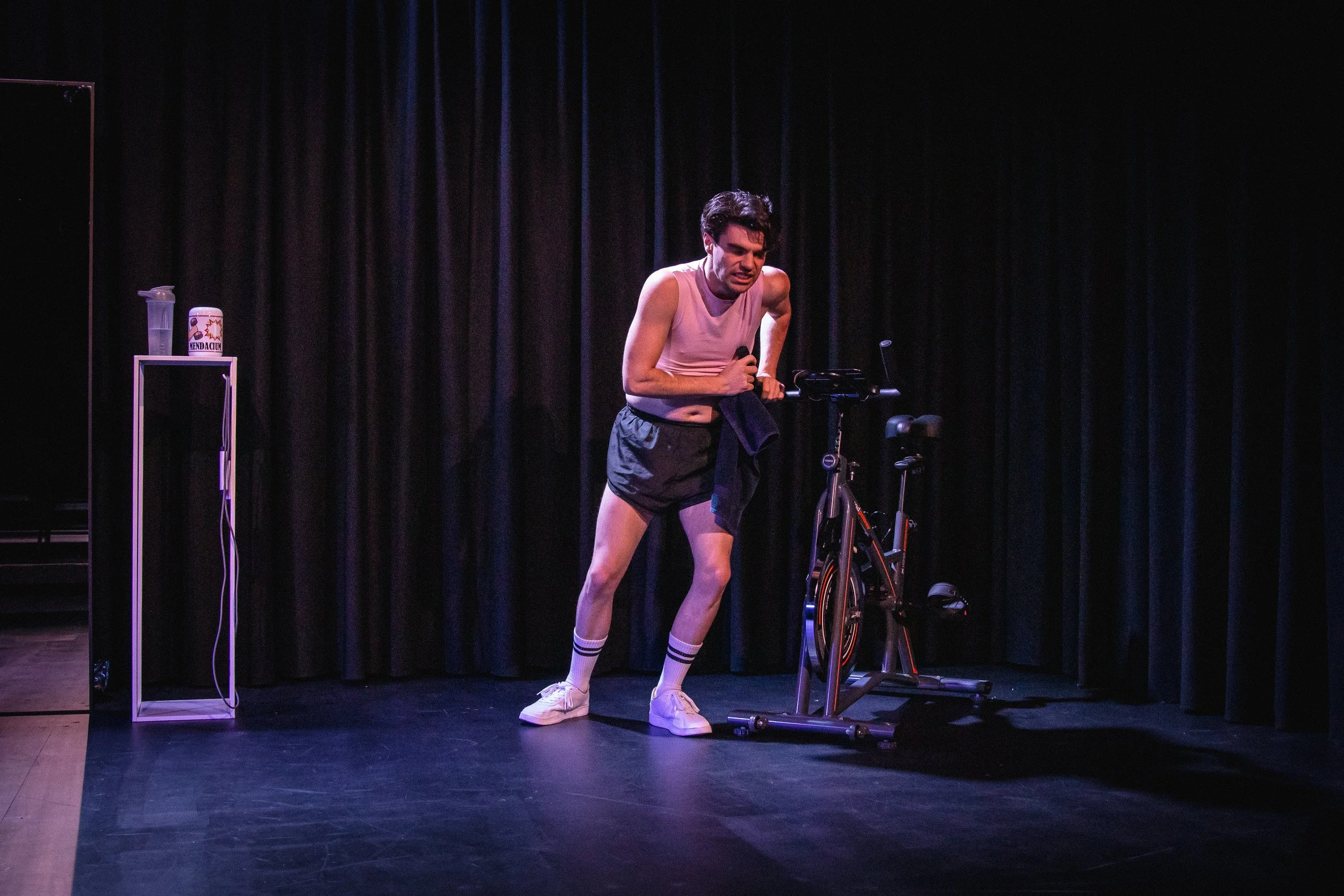 A man in athletic clothing appears exhausted, leaning on an exercise bike on a dark stage with black curtains, holding a microphone in his right hand.