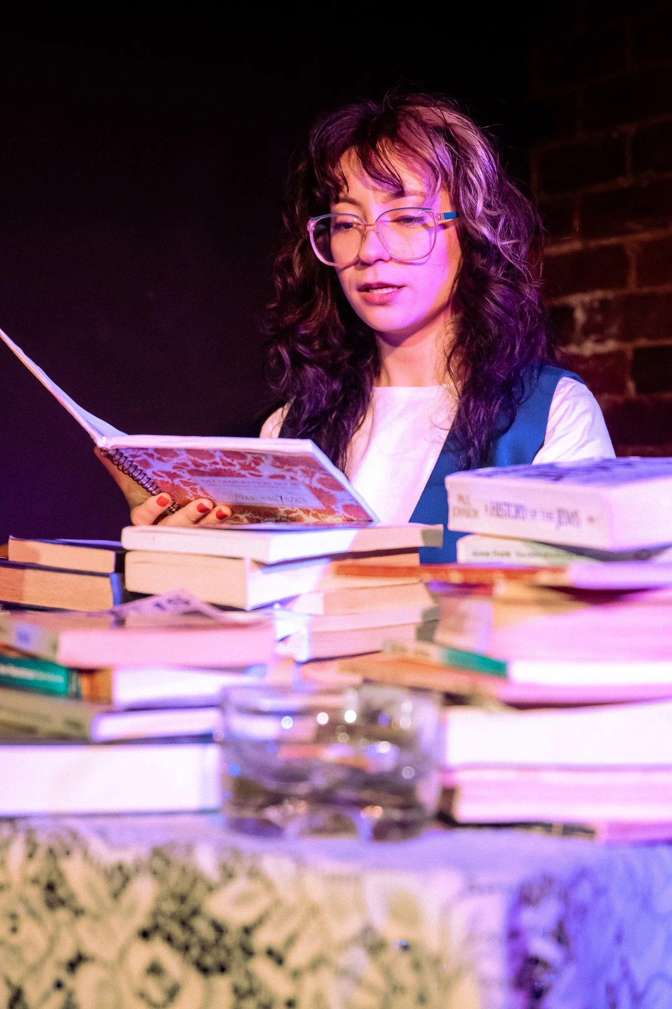 A woman with curly dark hair, wearing glasses and a white shirt, is reading a book at a table filled with stacks of books and a glass of water in a dimly lit room.