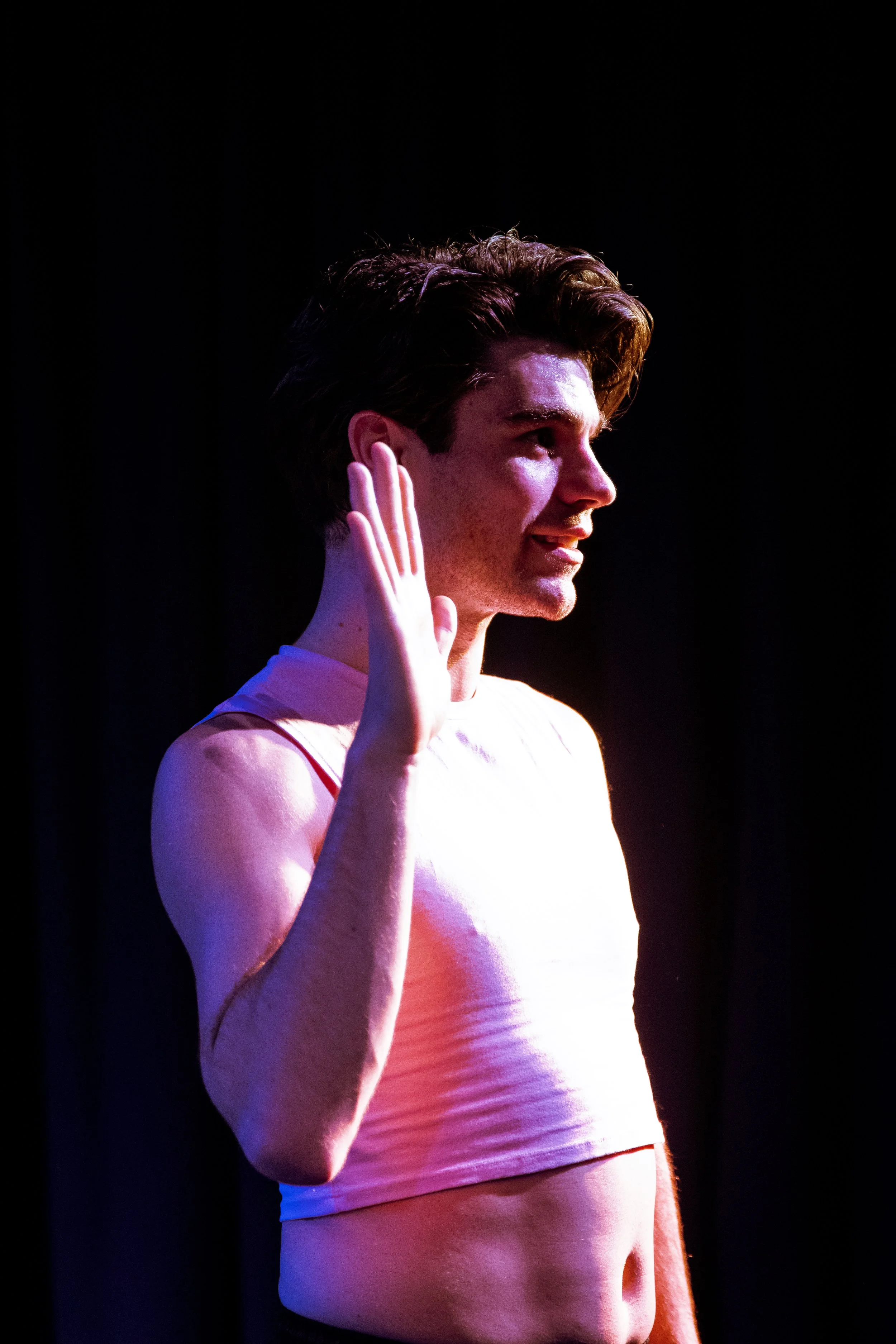 Young man with dark hair, wearing a sleeveless top, raising his right hand in a salute, standing against a dark background with stage lighting.