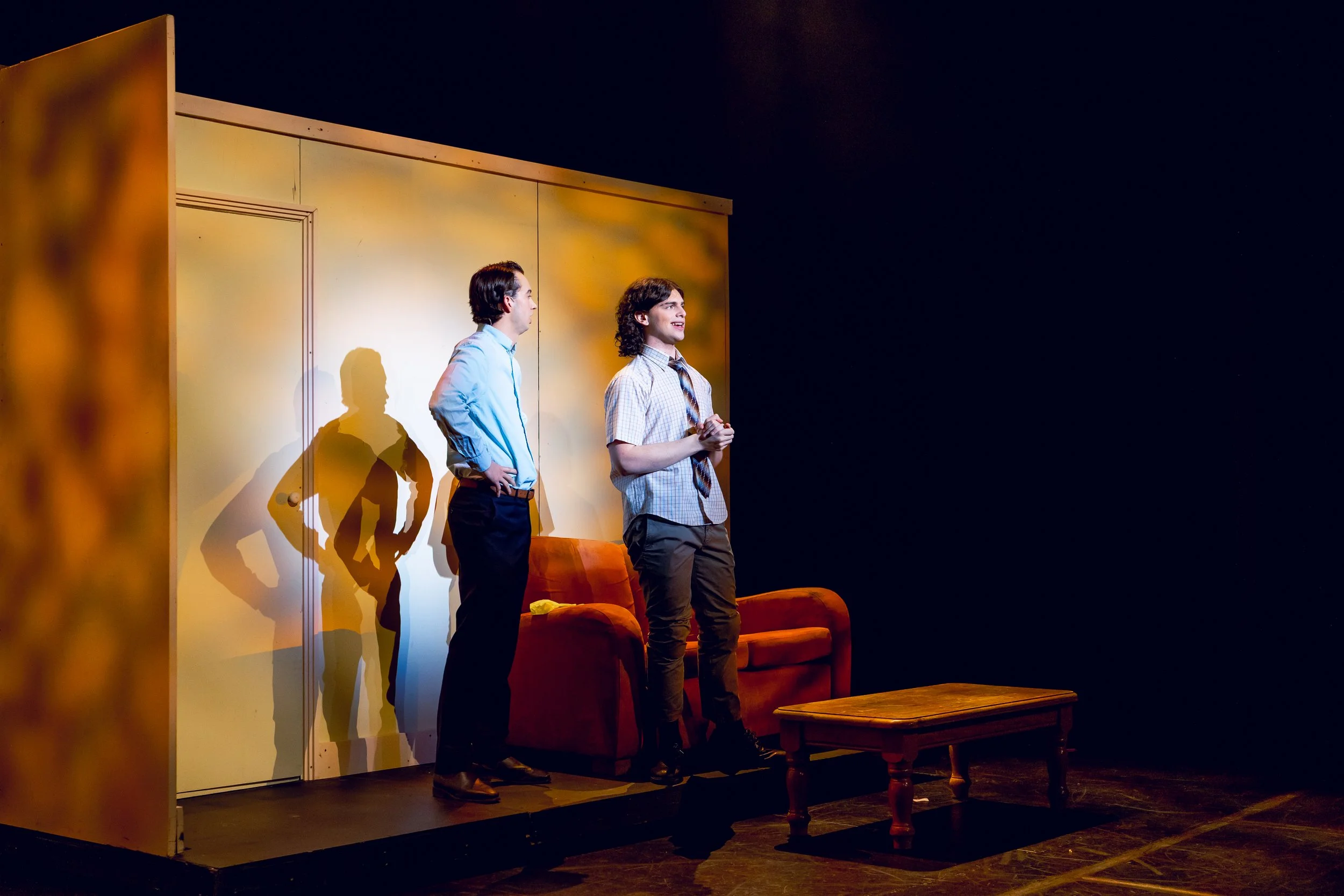Two men on stage in a theater, with shadows cast on the wall behind them, one with hand on hip and the other clasping hands, near a sofa and a wooden table.