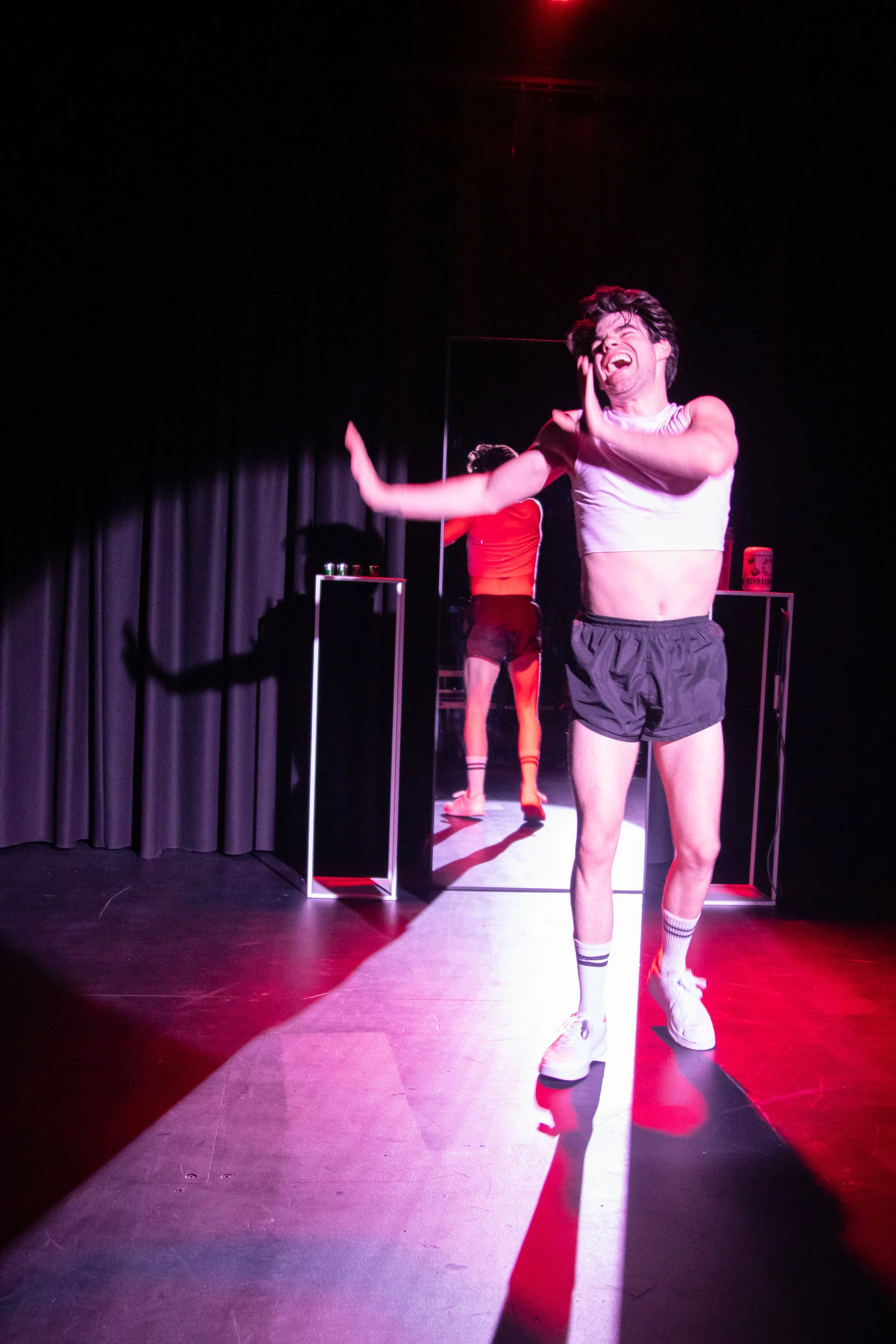 A man on stage wearing a white crop top, black shorts, and white sneakers, smiling and gesturing with his arms in front of a mirror with a woman in a red top and black shorts reflected behind him.