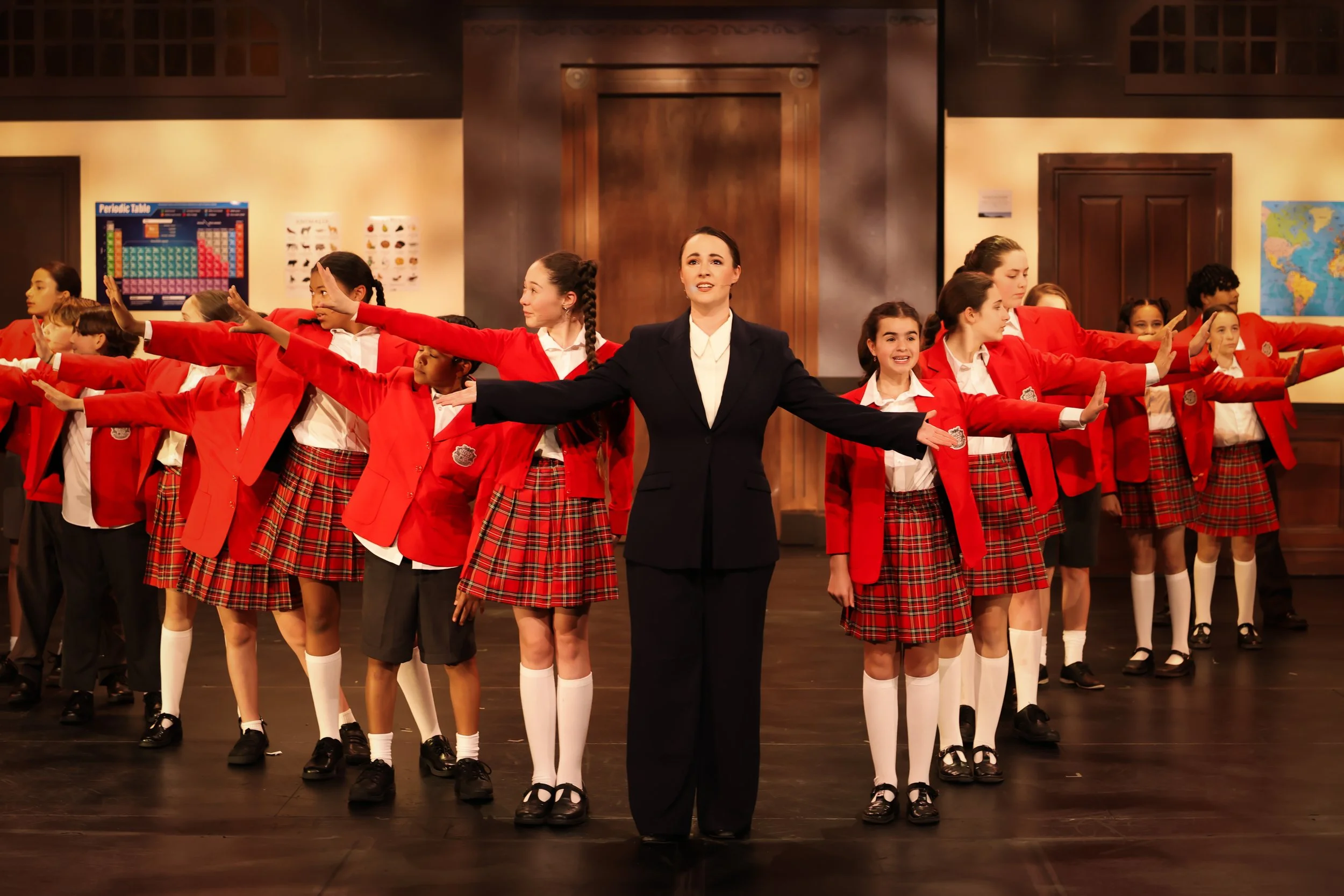 A group of school children in red blazers and plaid skirts standing with arms extended in a classroom setting, led by a woman in a dark suit.