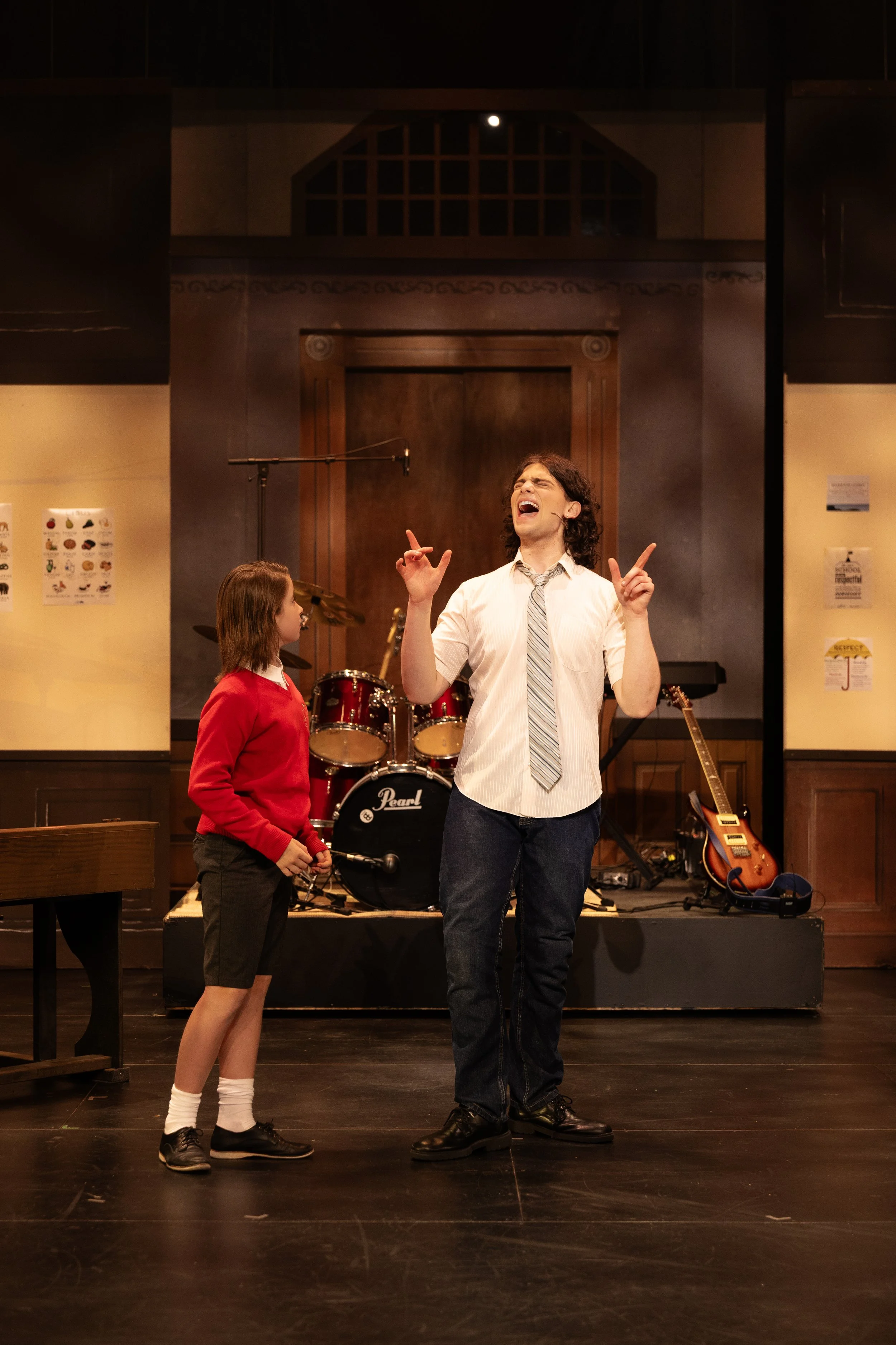 A scene from a school performance with a male actor singing on stage with a young girl watching, musical instruments like drums and guitar in the background.