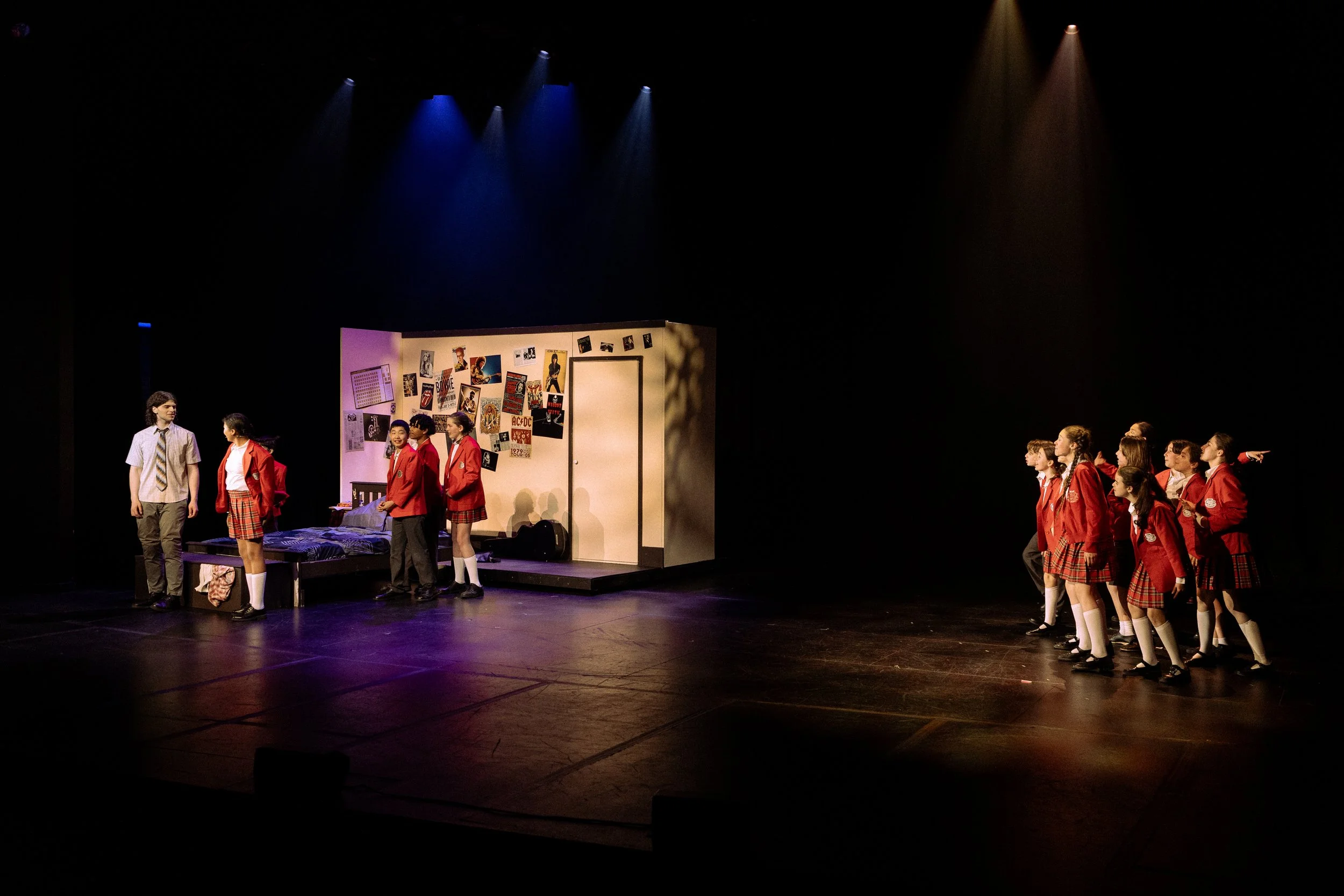 School students performing a play on stage with a bedroom set and wall decorated with posters and photos.