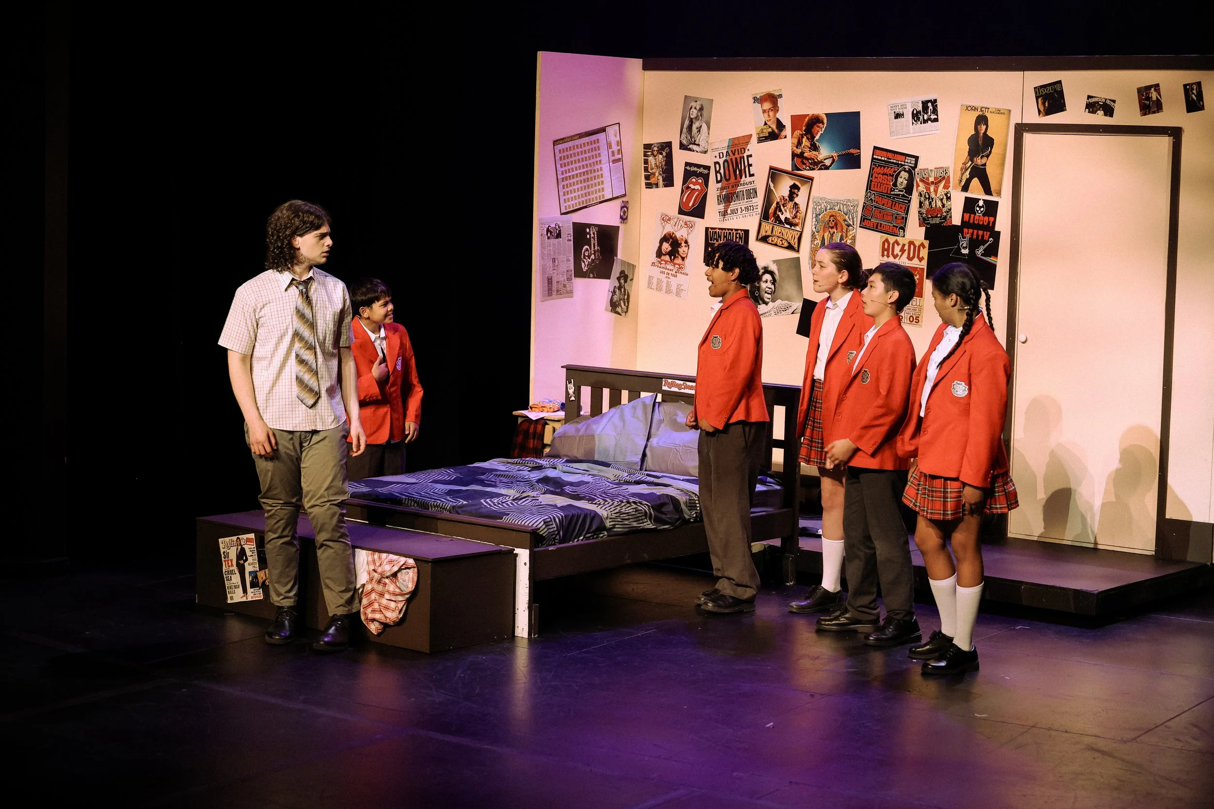Six actors on stage in a school-themed play, with five children in red blazers and one adult male in casual clothes, standing in a school bedroom set with posters of rock stars and musicians on the background wall.