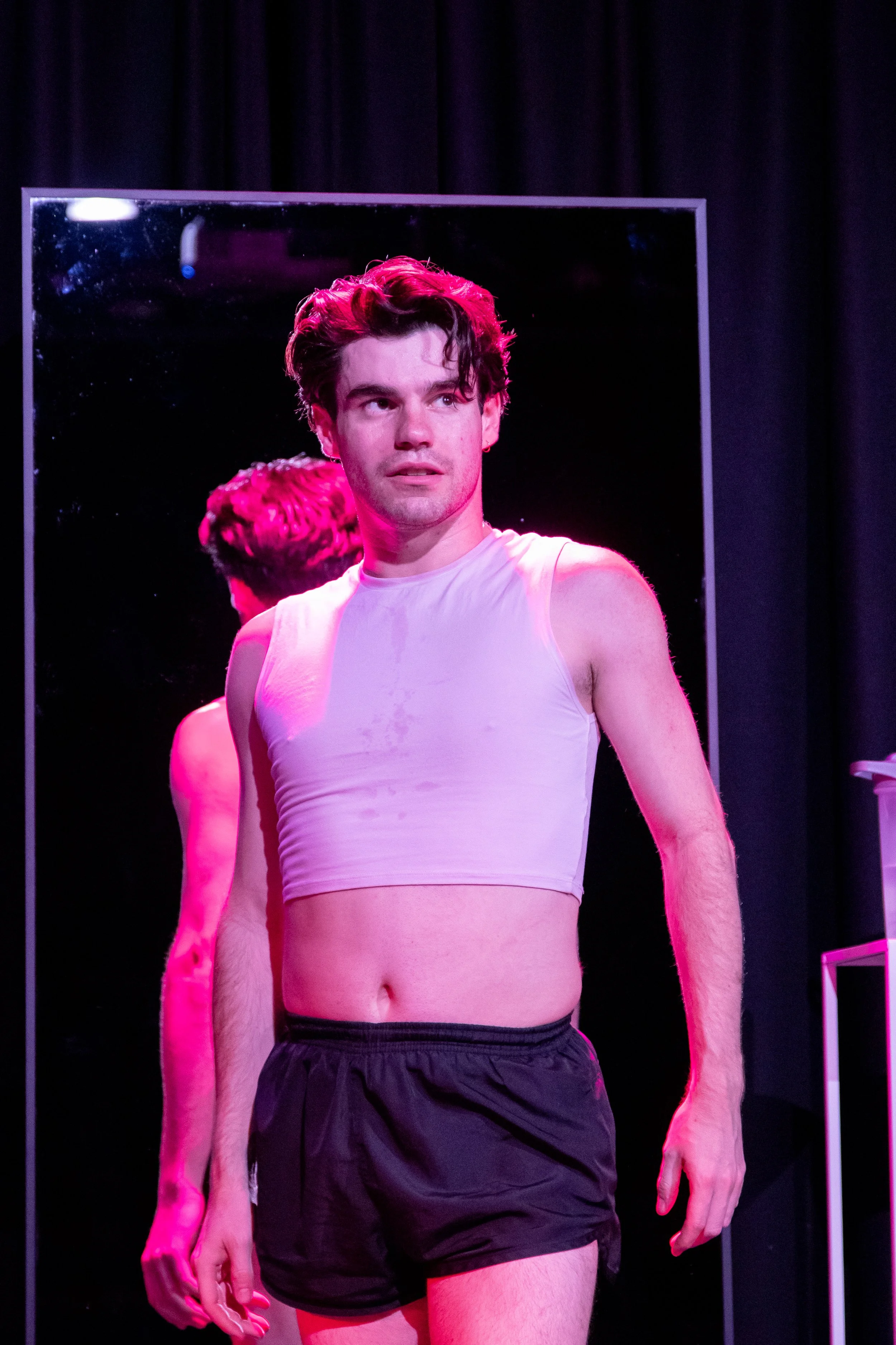 A man standing shirtless in front of a mirror on a stage, with a dark curtain behind him, wearing black shorts.
