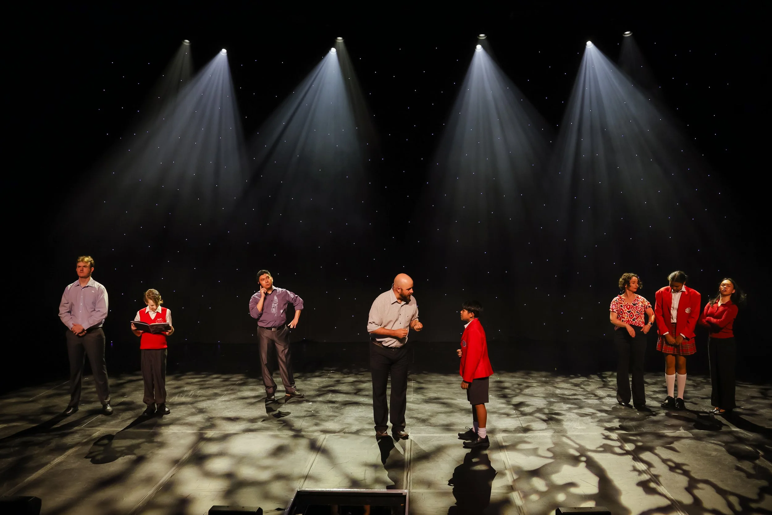 The image depicts a stage scene with seven people, including children and adults, under spotlights against a starry black background, appearing to be in a theatrical or musical performance.