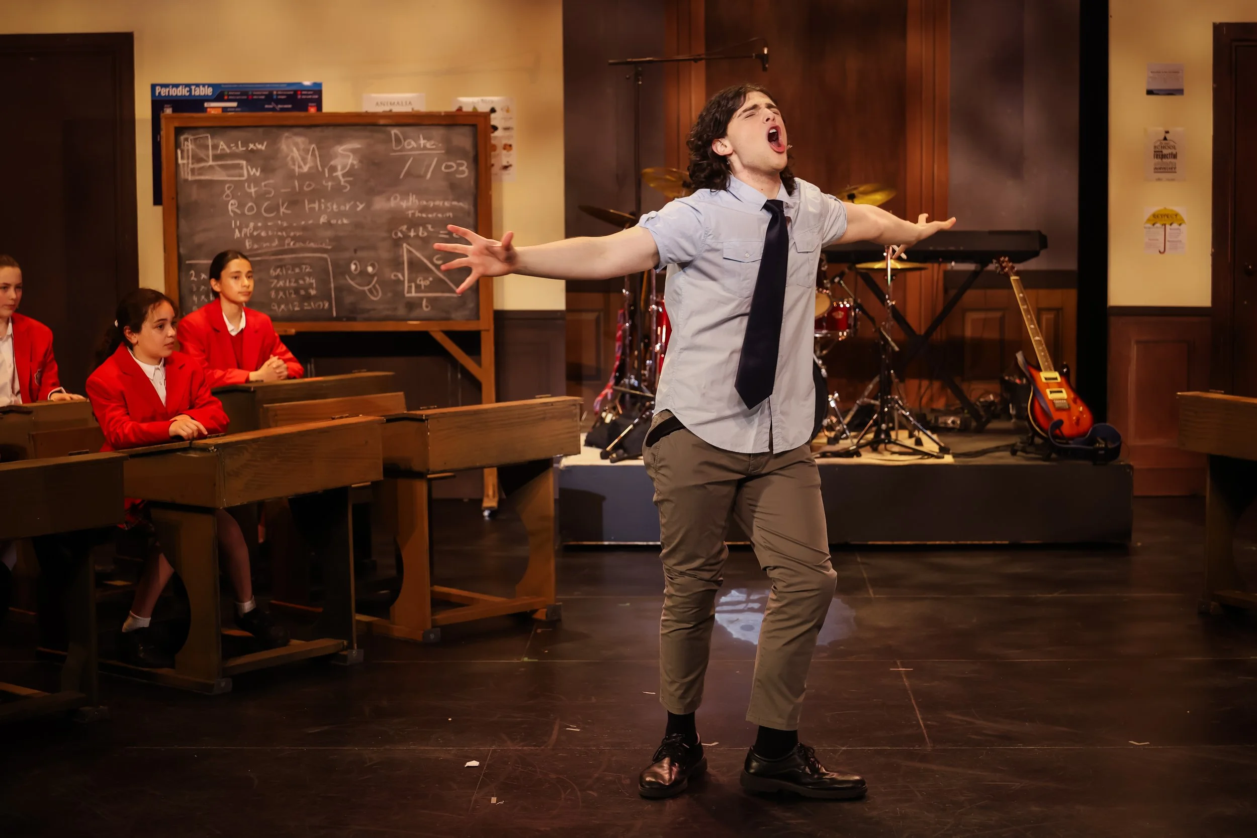 A young man with long curly hair wearing a light blue shirt, black tie, and khaki pants, singing passionately with arms outstretched in a classroom or performance space, with students in red uniforms sitting at desks and musical instruments on stage 